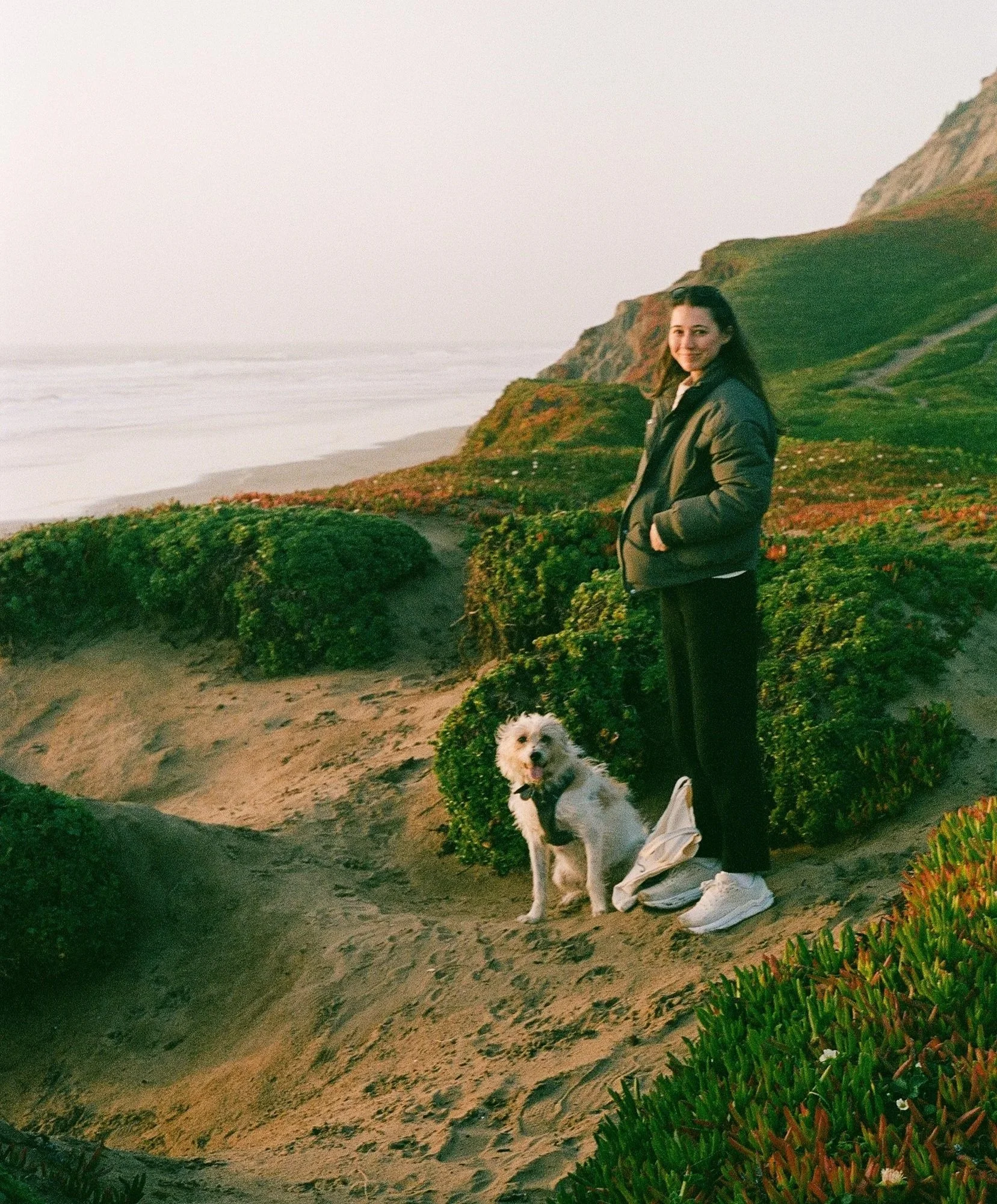A woman and a small white dog with curly fur standing on a sandy trail near green bushes and coastal cliffs at sunset.