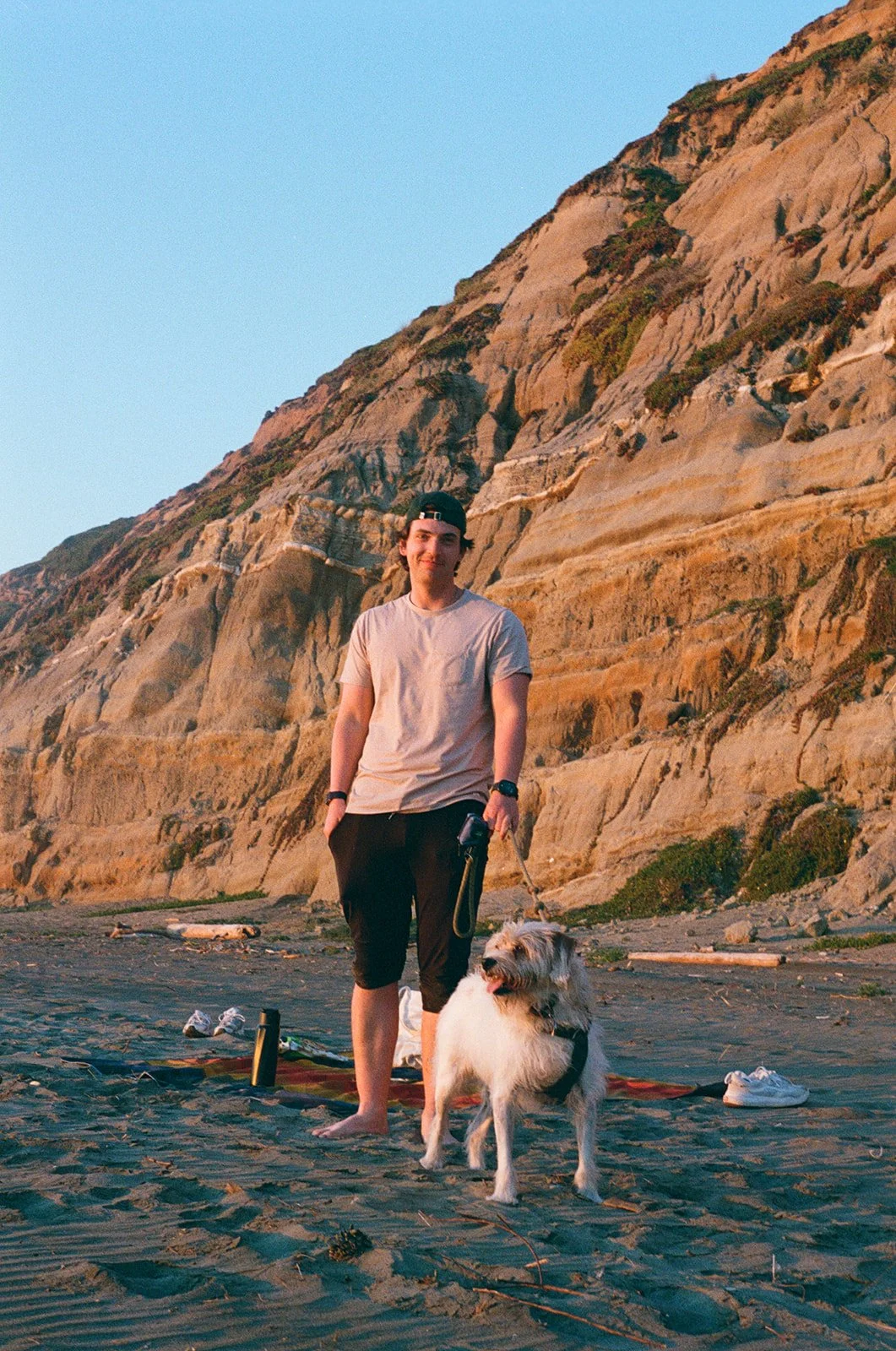 A young man standing on a sandy beach with a dog, with a large rocky cliff in the background at sunset.