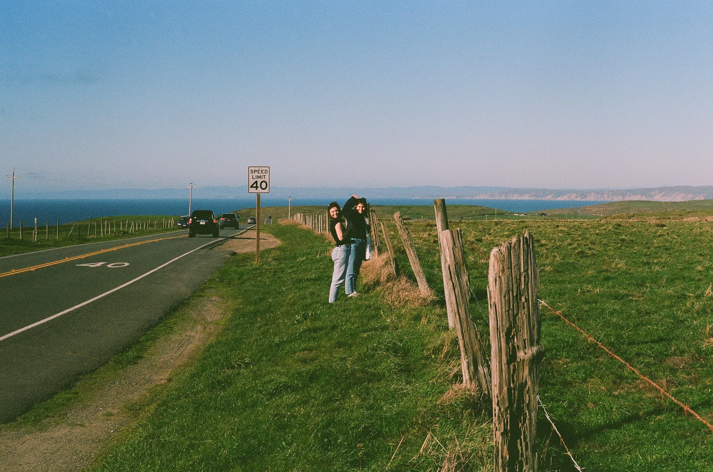 Two women standing near a wooden fence along a rural road with a speed limit sign of 40, overlooking a coastline with hills and water in the background.
