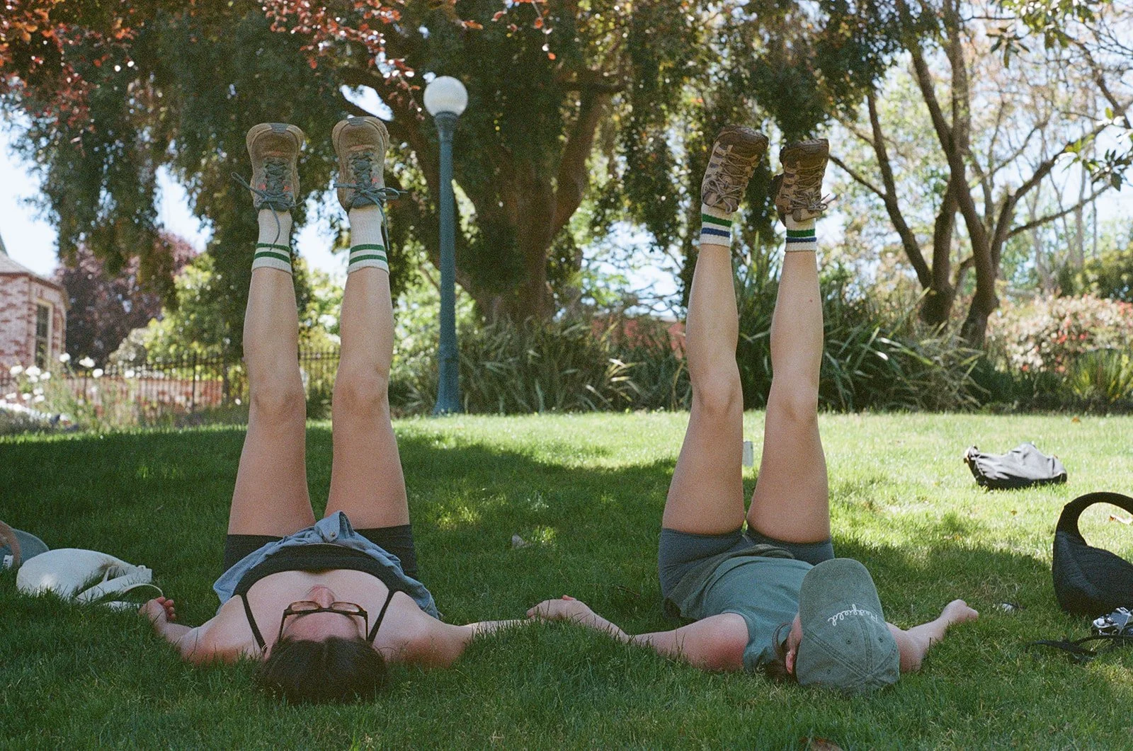 Two children lying on grass with their legs raised vertical in a park, wearing knee-high socks and sneakers, surrounded by trees and foliage.