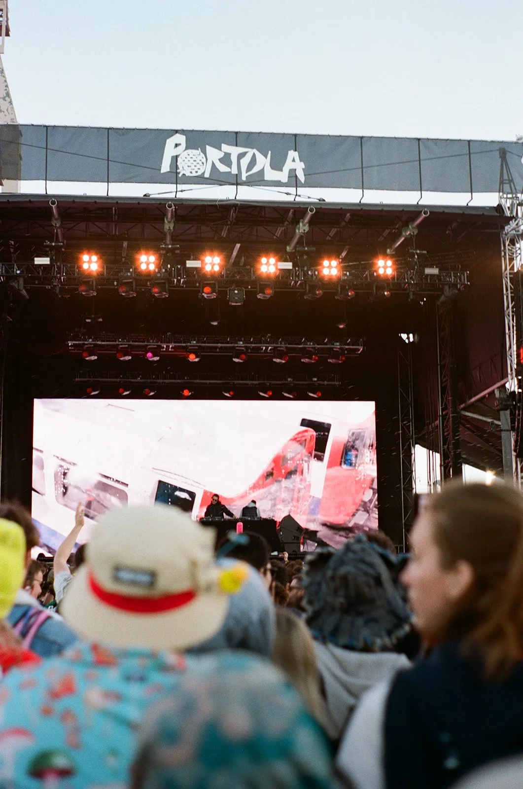Crowd attending an outdoor music festival or concert at the Portola festival, with a stage, DJ booth, and a large digital screen showing visuals.