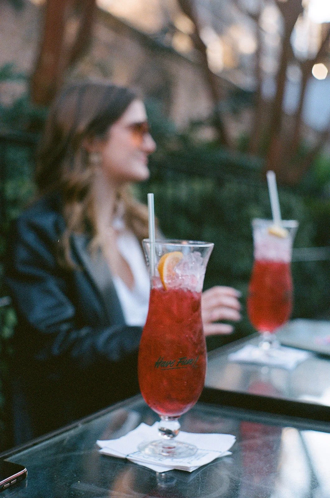 A woman sitting at a table outdoors with two tall glasses of red drinks garnished with lemon slices and ice, one in the foreground and one in the background, blurred. She is wearing sunglasses and a dark blazer over a white shirt, with trees and a wo