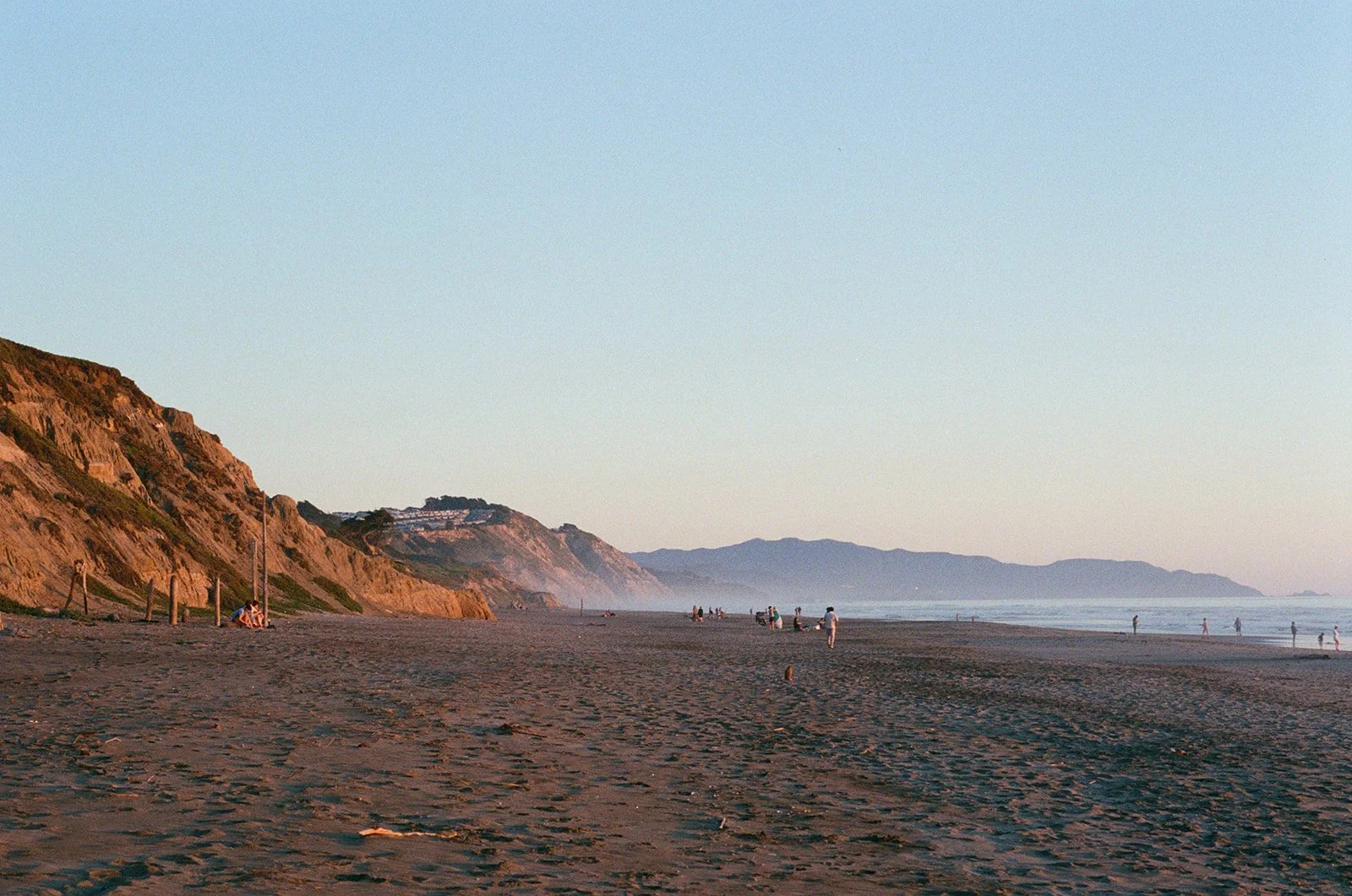 People enjoying a sandy beach with cliffs on the left and ocean waves in the background at sunset.