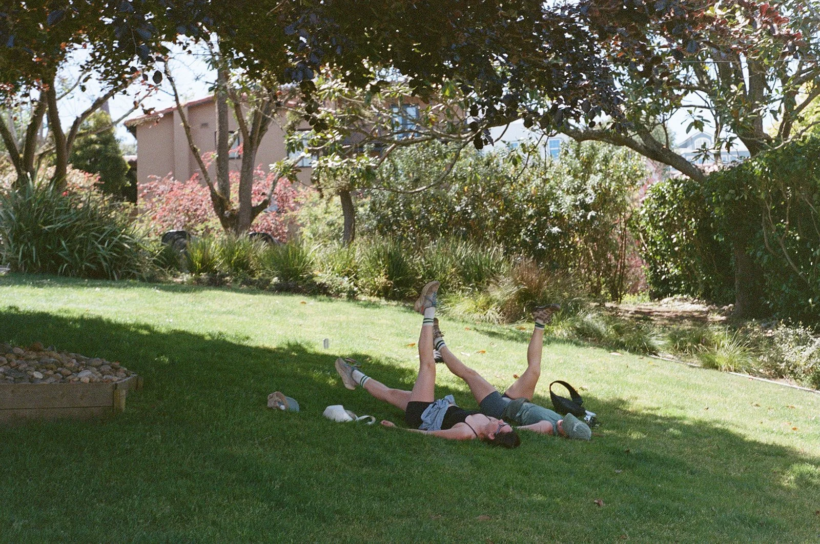 Two children wearing summer clothes play on grass in a sunny park, with trees and bushes in the background.