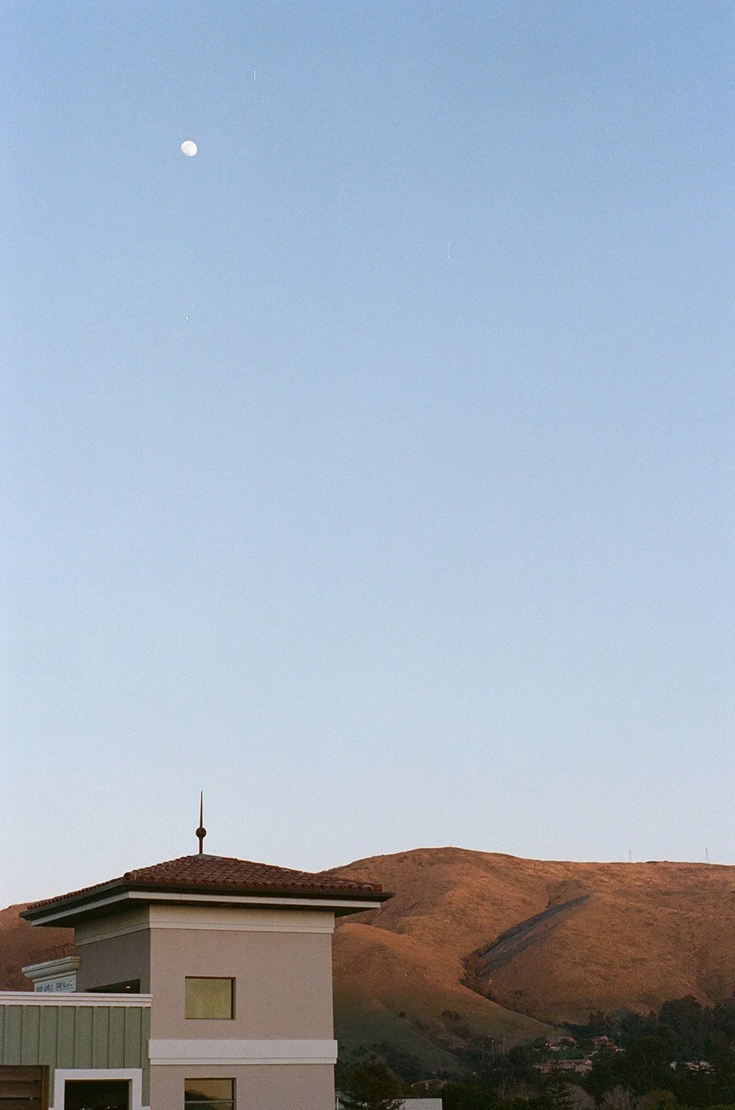 A house with a red-tiled roof and a small window on the upper floor, set against a backdrop of rolling hills with dry grass, under a clear sky with the moon visible.