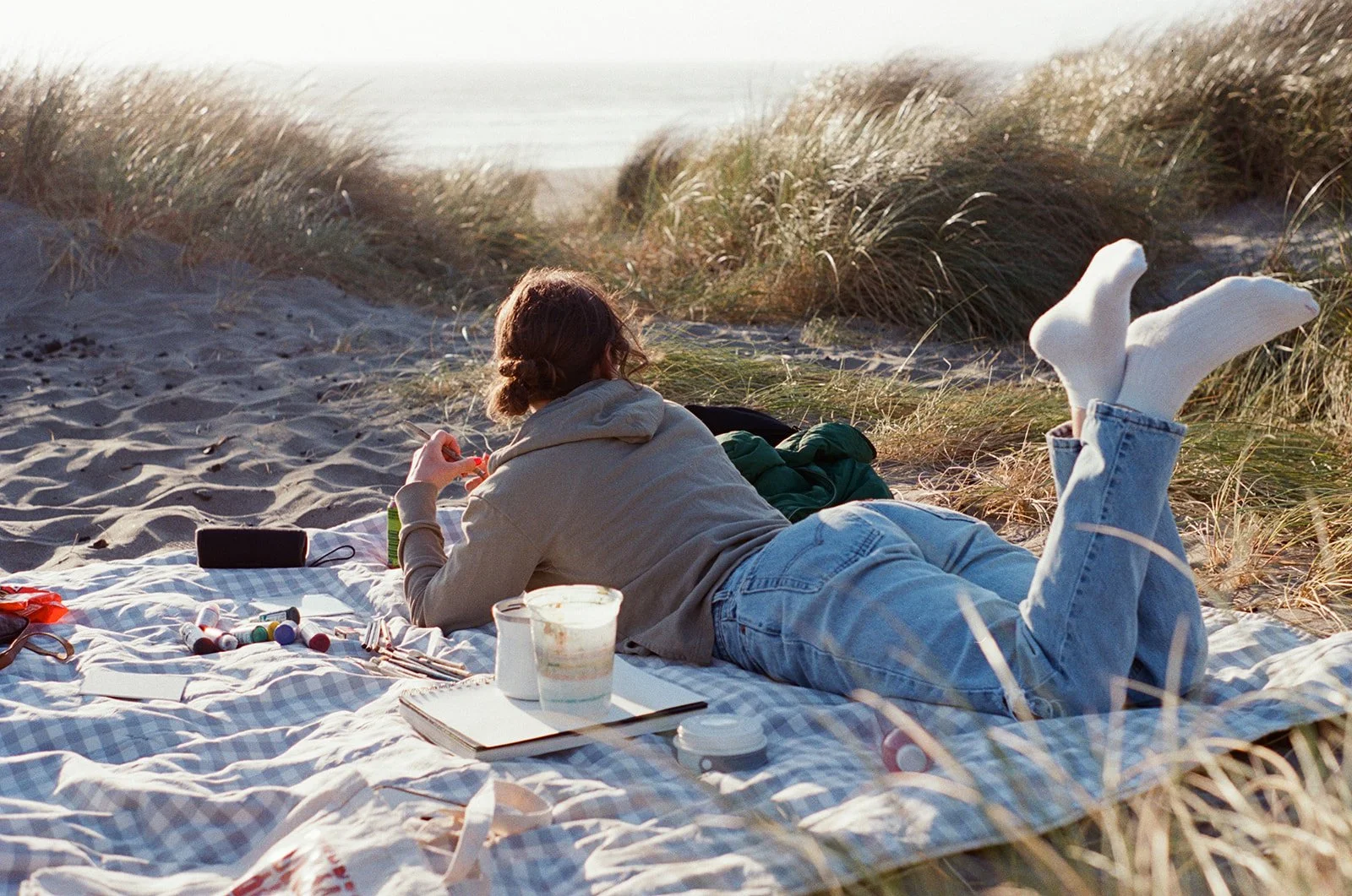 Person lying on a checkered blanket on a sandy beach with tall grass in the background. They are wearing light-colored jeans and white socks, with their legs crossed in the air. Items on the blanket include paint supplies, a black speaker, a book, an