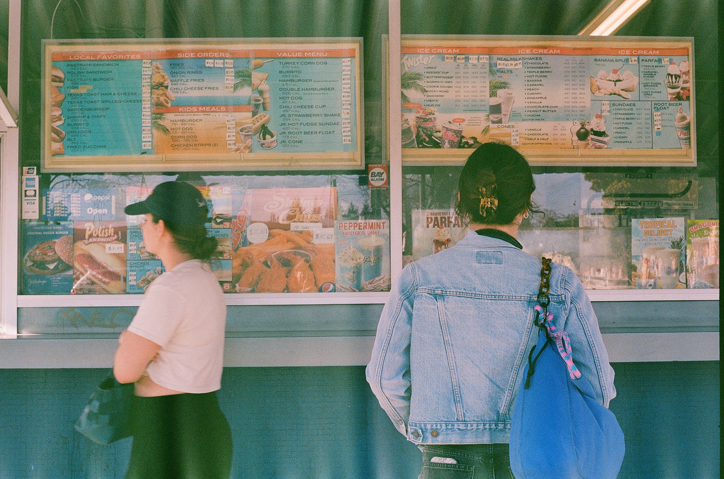 Two women stand outside a fast food restaurant window, looking at the menu; one wears a black cap and white shirt, the other has curly hair in a denim jacket and carries a colorful backpack.