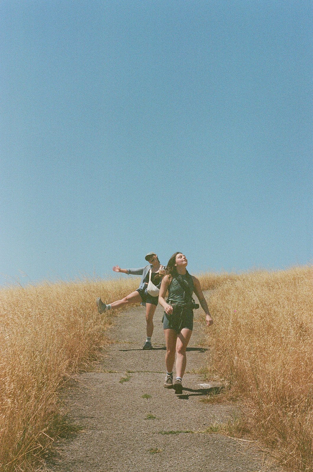 Two young women are walking along a paved path through a field of tall, golden grass. One woman is in front, looking up with her eyes closed. The other woman is behind, balancing on one leg with her arm outstretched. The sky above is clear and blue.