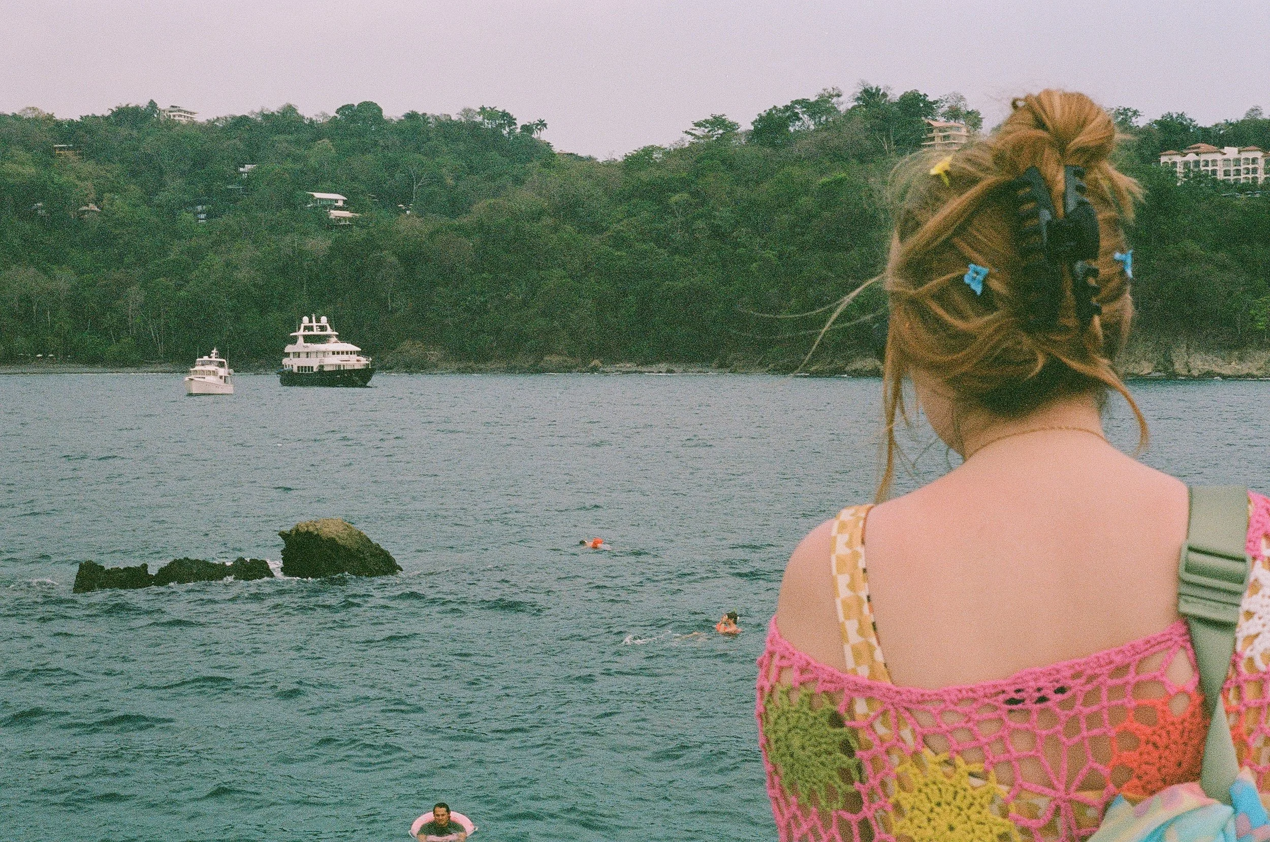 A woman with long, light brown hair in a ponytail, wearing a colorful crochet top with pink, yellow, and green patterns, stands near the water with her back to the camera. In the water, there are several people swimming, and a few boats are anchored 