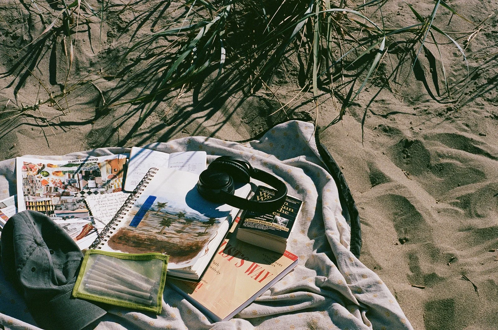 A towel spread on sandy ground with magazines, a notebook, headphones, a book, a gray cap, and a small transparent green container, near some grass and footprints.