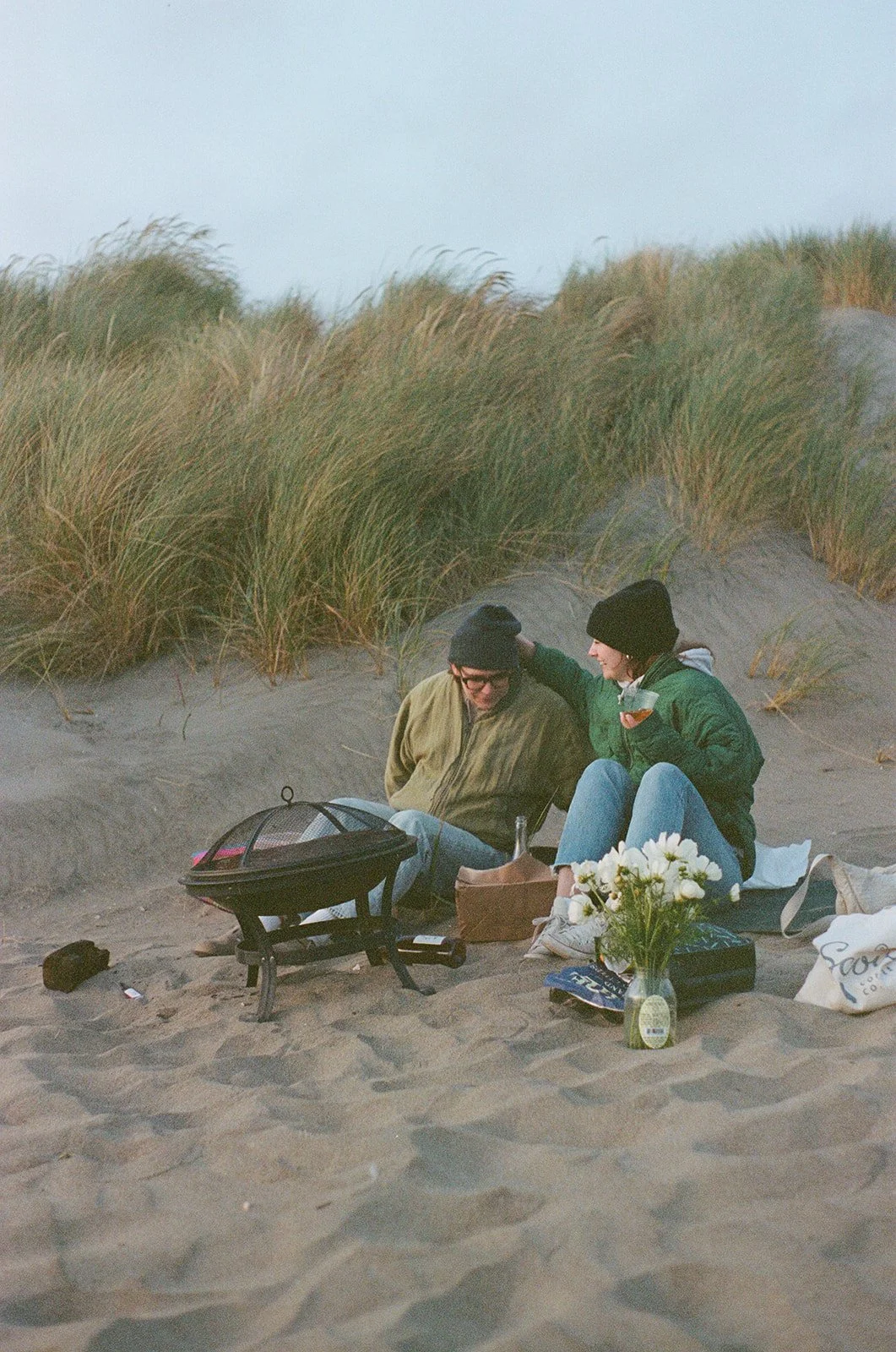 Two people sitting on the sand by grassy dunes, having a picnic with a small grill, flowers in a vase, and drinks, wearing jackets and beanies.