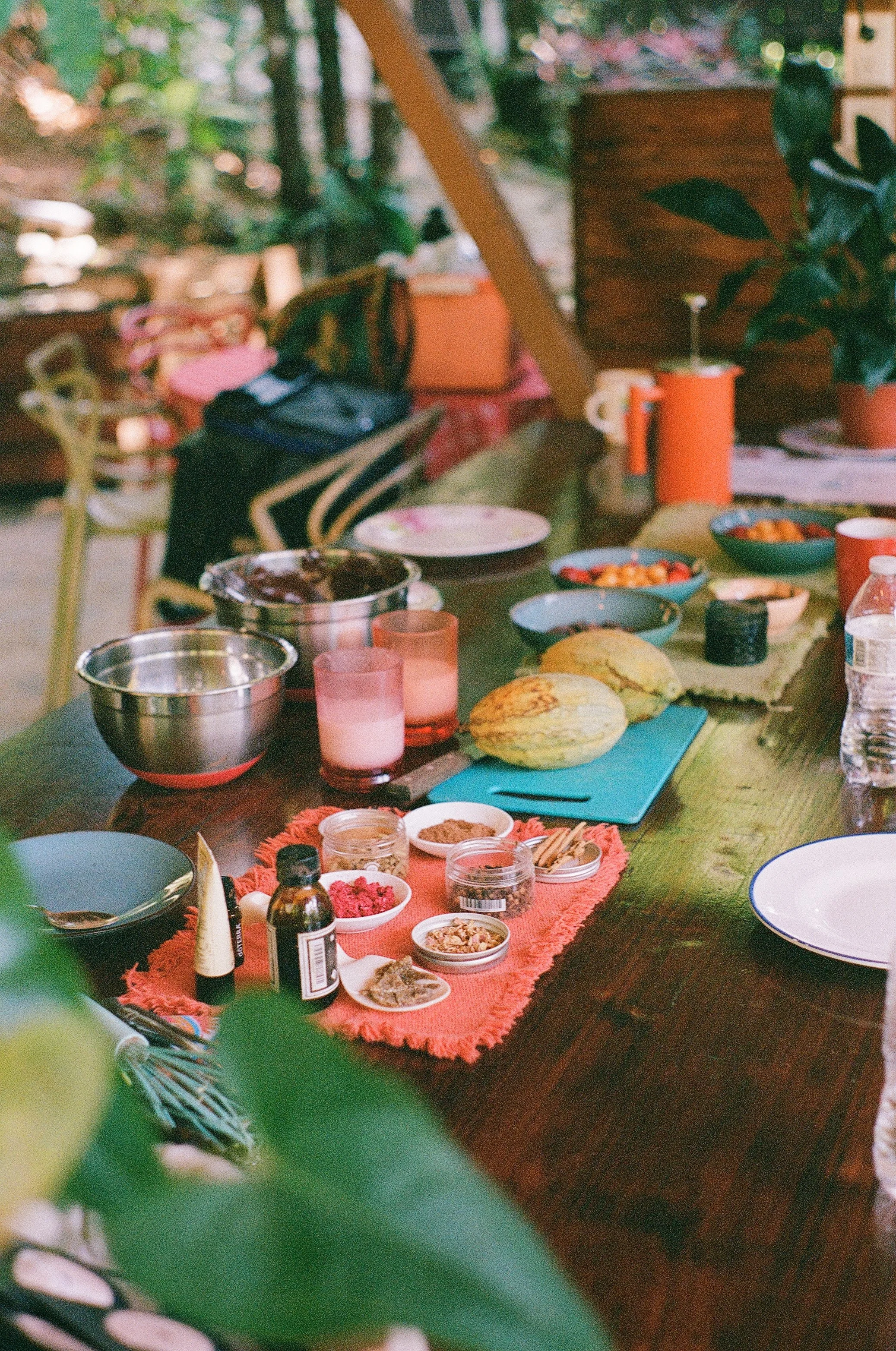 A table set for a meal with various bowls of food, plates, spices, and drinks on a green tablecloth. In the background, there are chairs and a wooden structure, likely part of a kitchen or outdoor area.