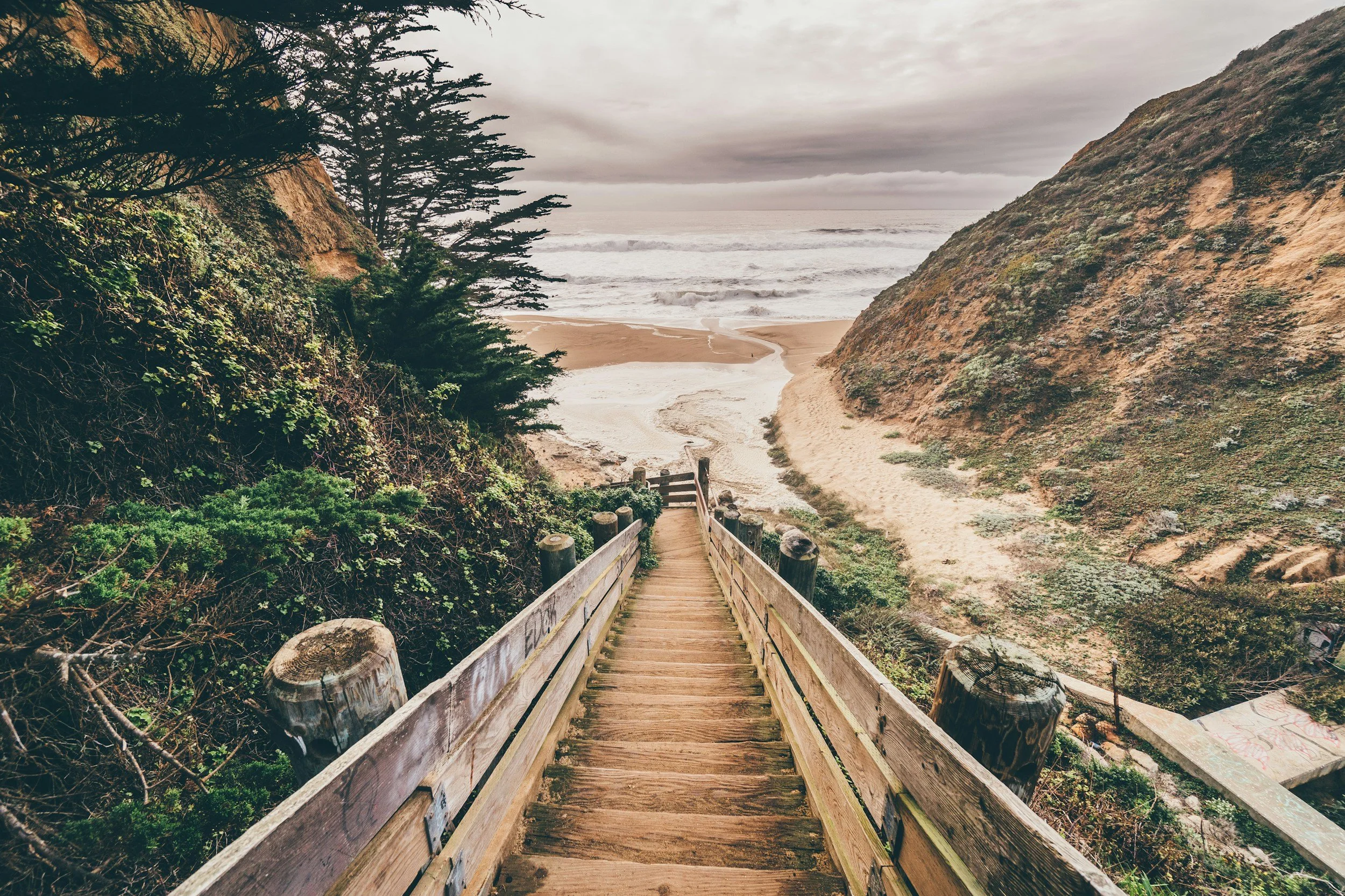 A wooden staircase leading down through lush greenery to a sandy beach with waves and a cloudy sky.