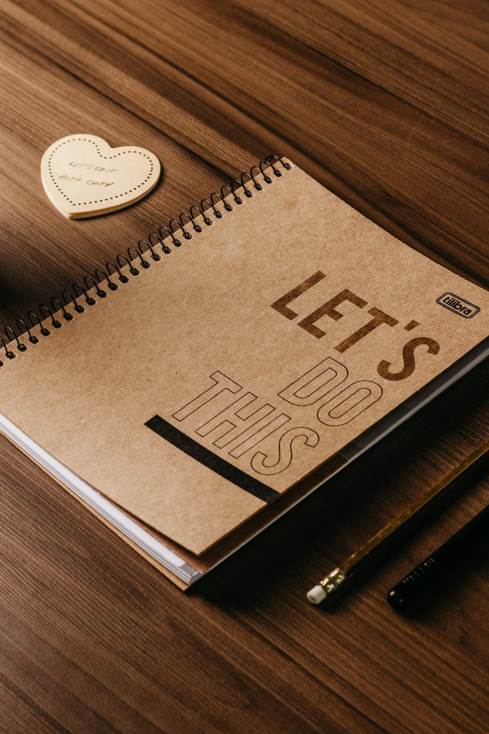 A notebook with the words "LET'S DO THIS" on the cover, a pen, a pencil, and a heart-shaped note that says "WISH YOU A WON COW" on a wooden surface.