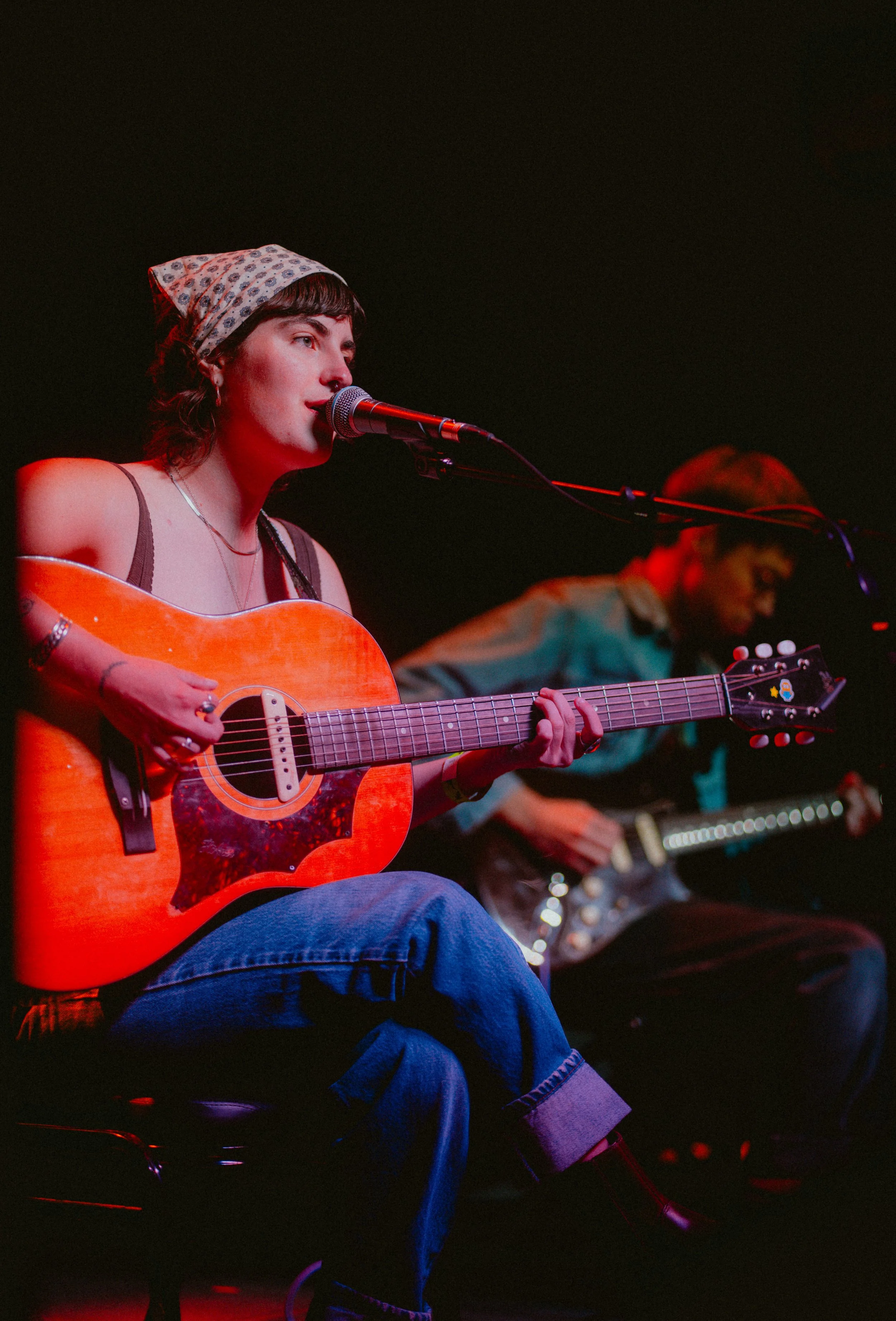 A female musician wearing a patterned headscarf and tank top, playing an orange acoustic guitar and singing into a microphone on stage, with another person playing a keyboard in the background.