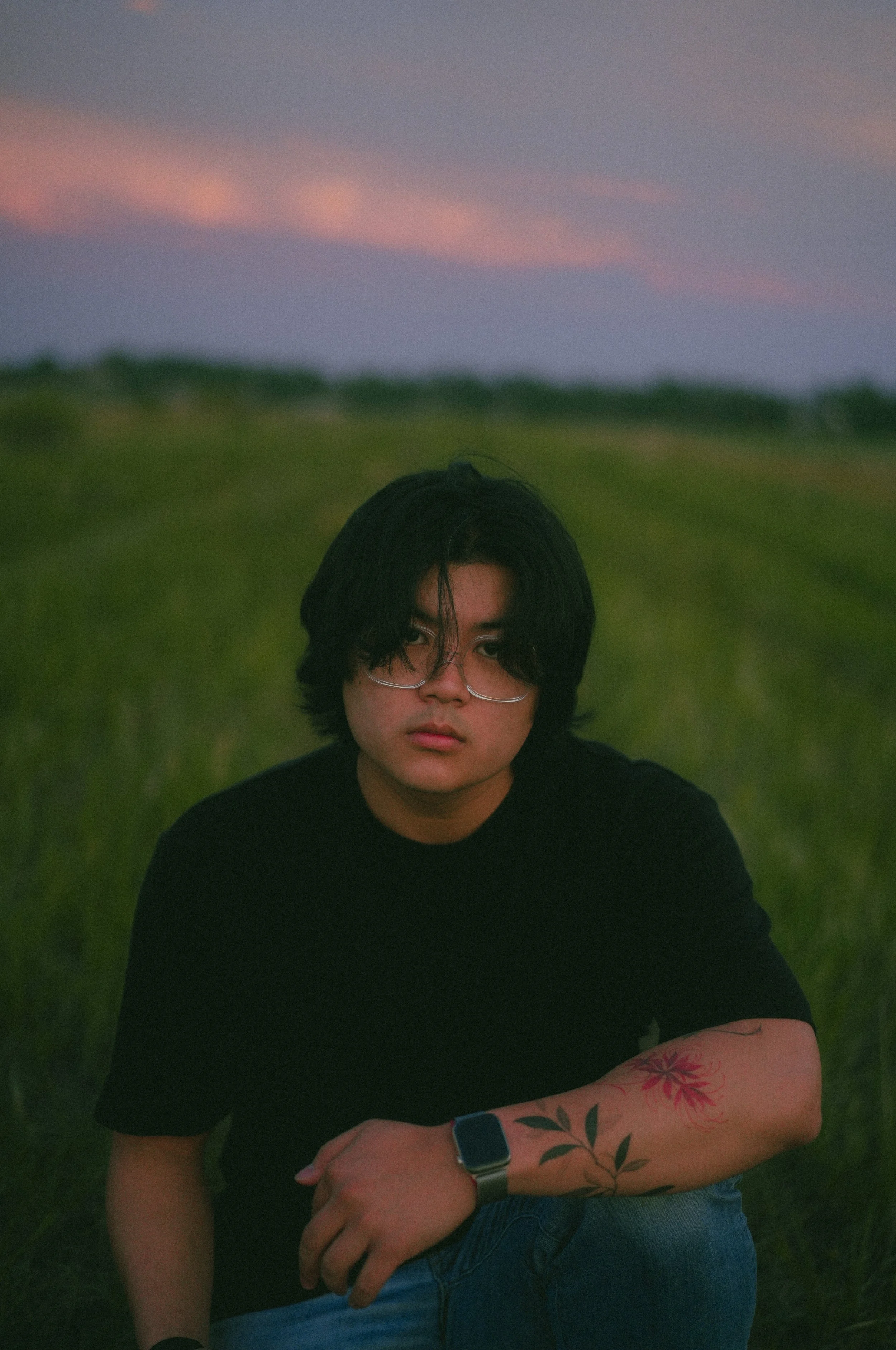 A young person with black hair and glasses sitting outdoors at sunset, wearing a black t-shirt with floral tattoos on one arm.