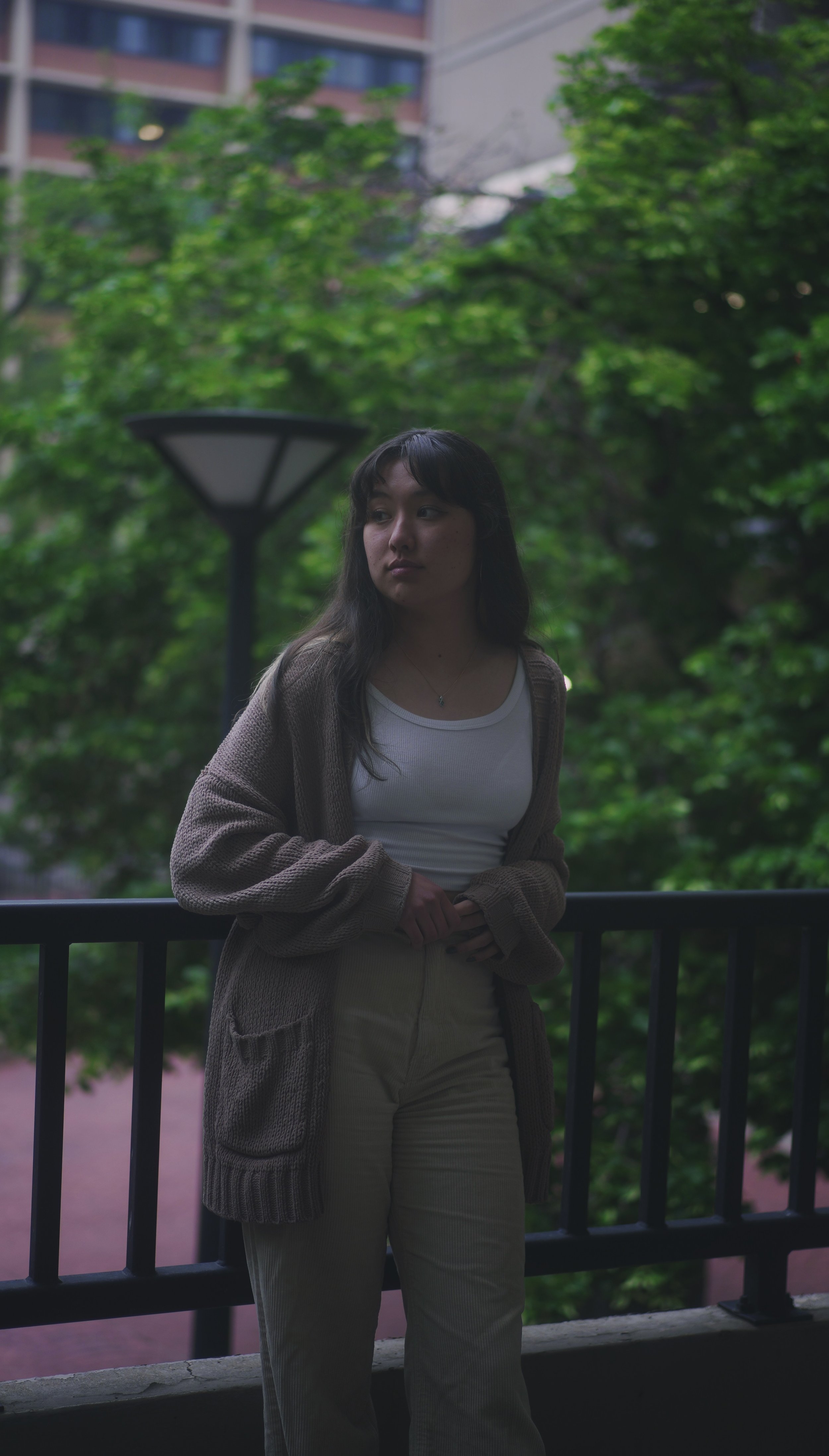 A young woman with dark hair stands outside on a balcony or terrace, leaning against a black railing. She is wearing a white top and beige pants, with a brown cardigan. Behind her, there is lush green foliage and a tall street lamp, with a background