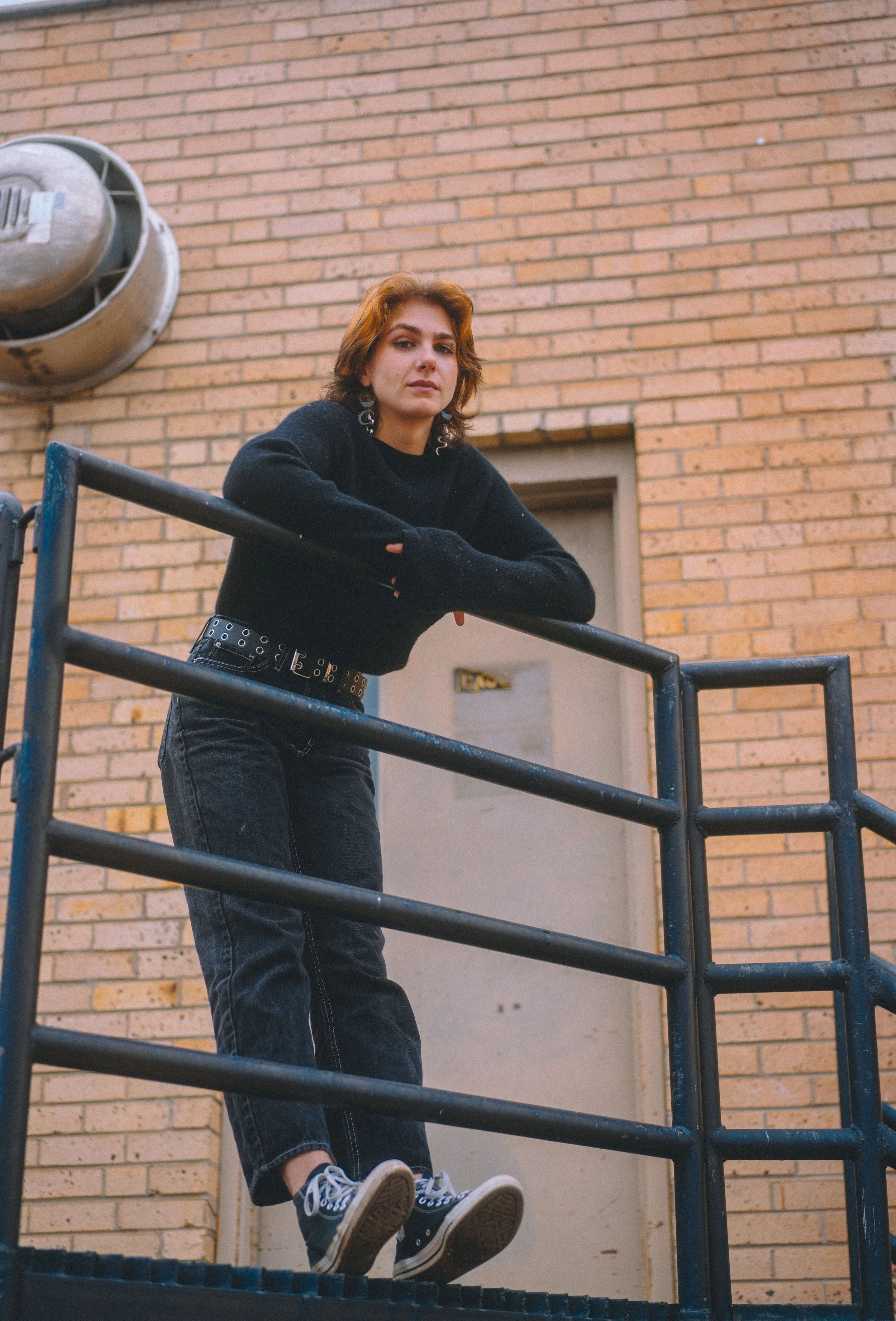 A young woman with shoulder-length reddish-brown hair leaning on a railing in front of a brick building.