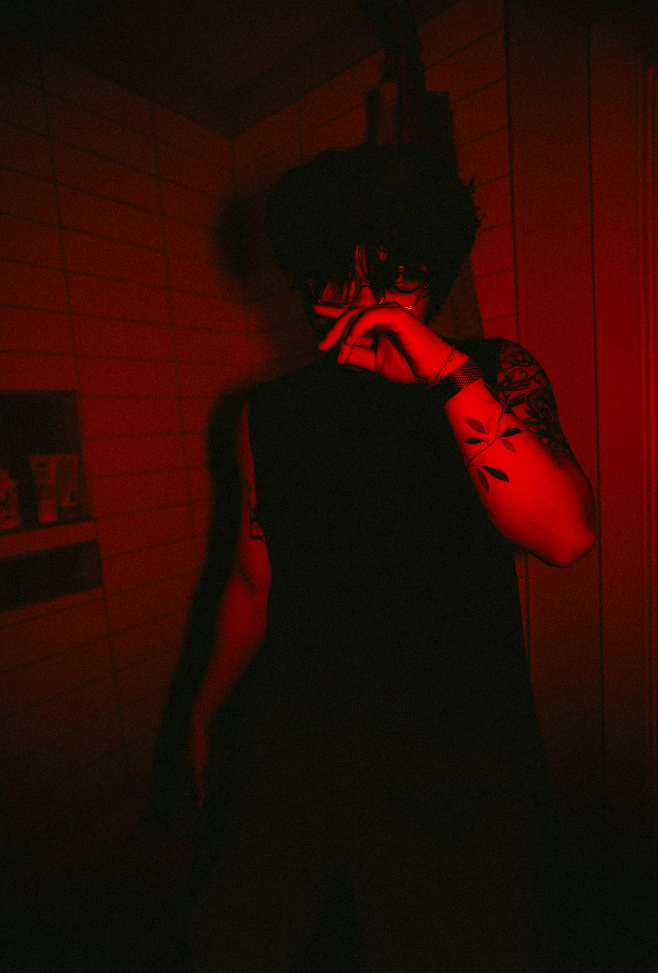 Person with curly hair and glasses posing in front of a tiled wall, red lighting, wearing a black top with a tattooed arm visible, hand near face.