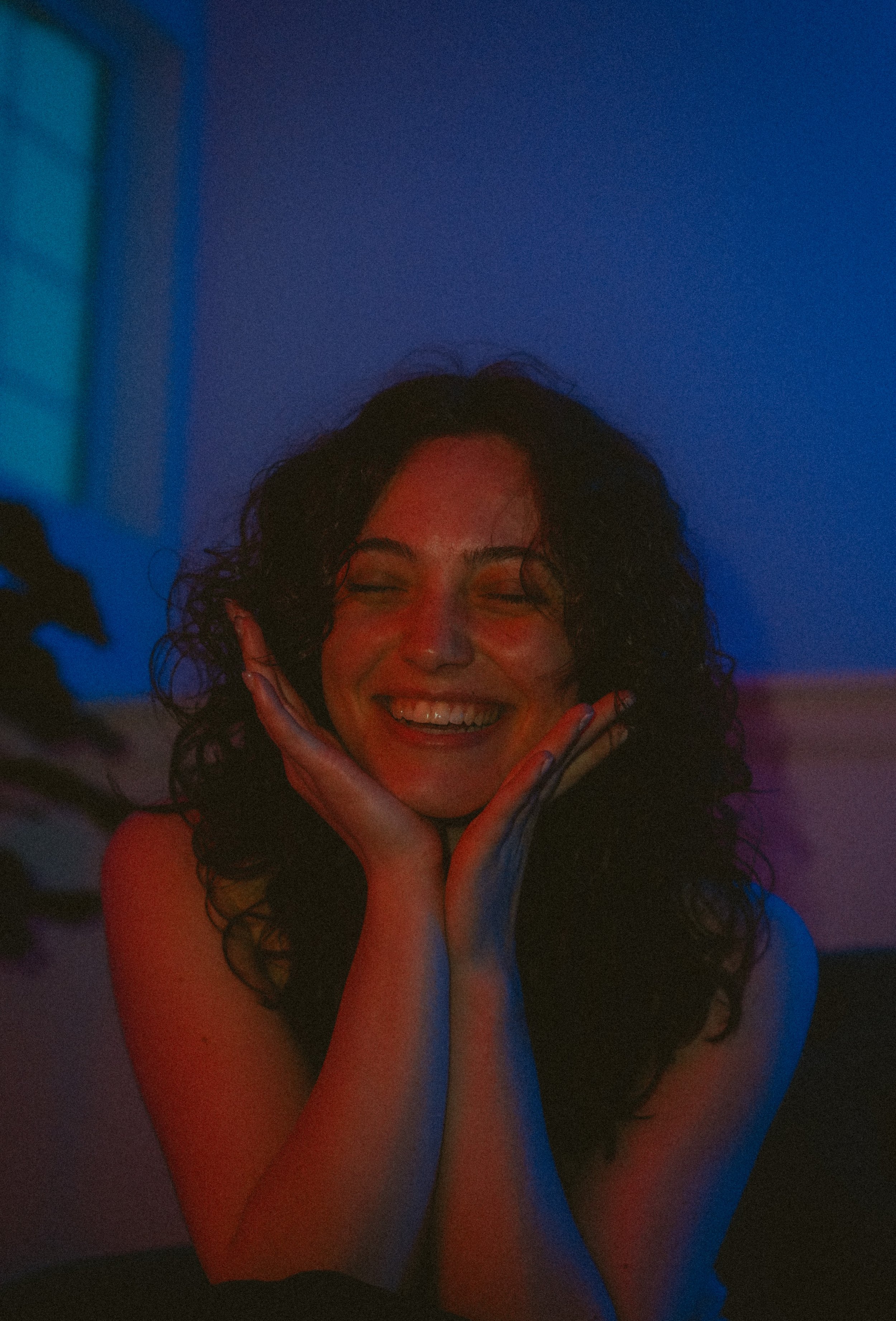 A woman with curly dark hair smiling with eyes closed, hands framing her face, in colorful lighting with blue and red hues.
