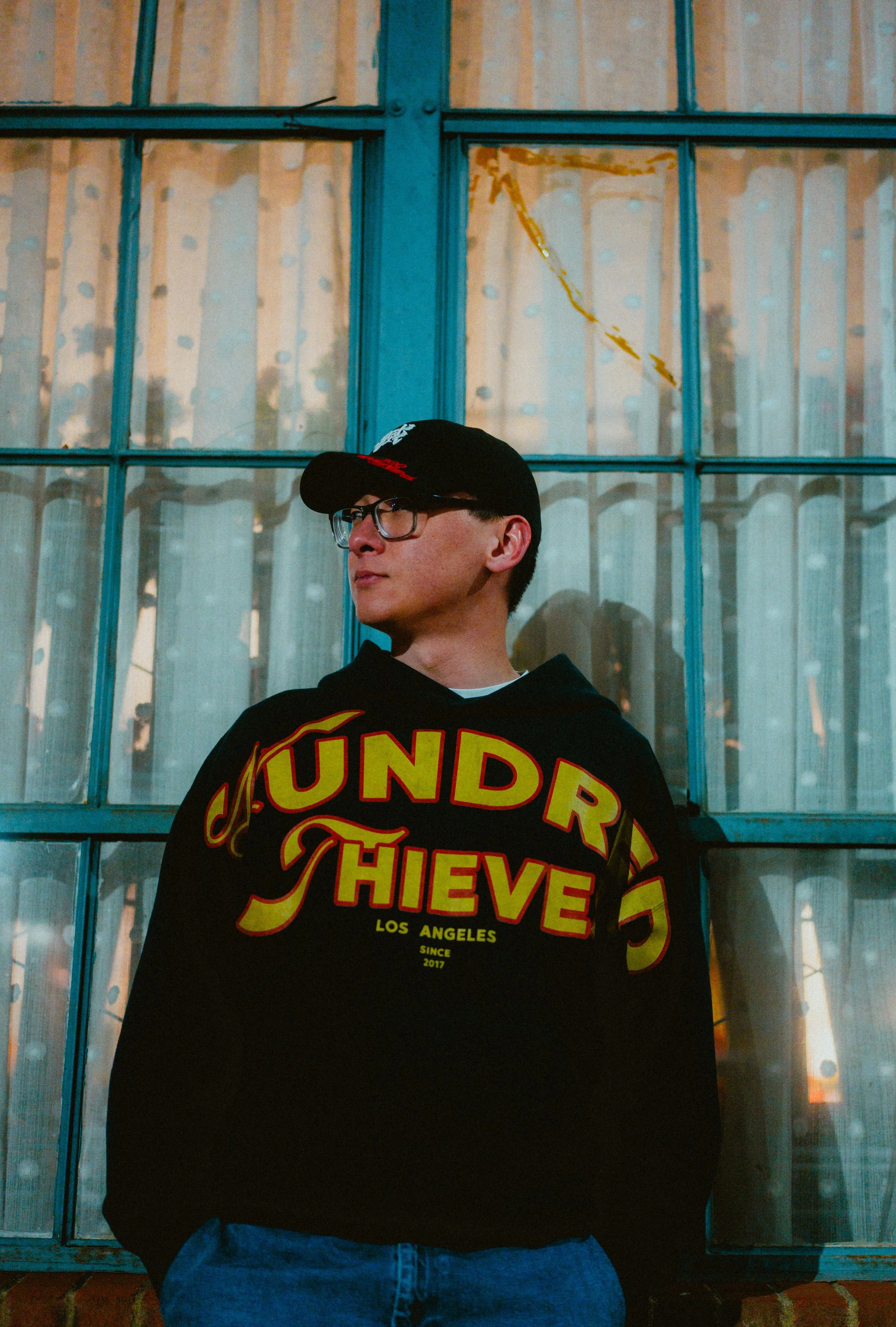 A young man wearing glasses, a black cap, and a black hoodie with colorful text stands in front of a large, old, blue-framed window with peeling paint. The window has multiple panes, some of which are foggy or reflective. The background behind the wi