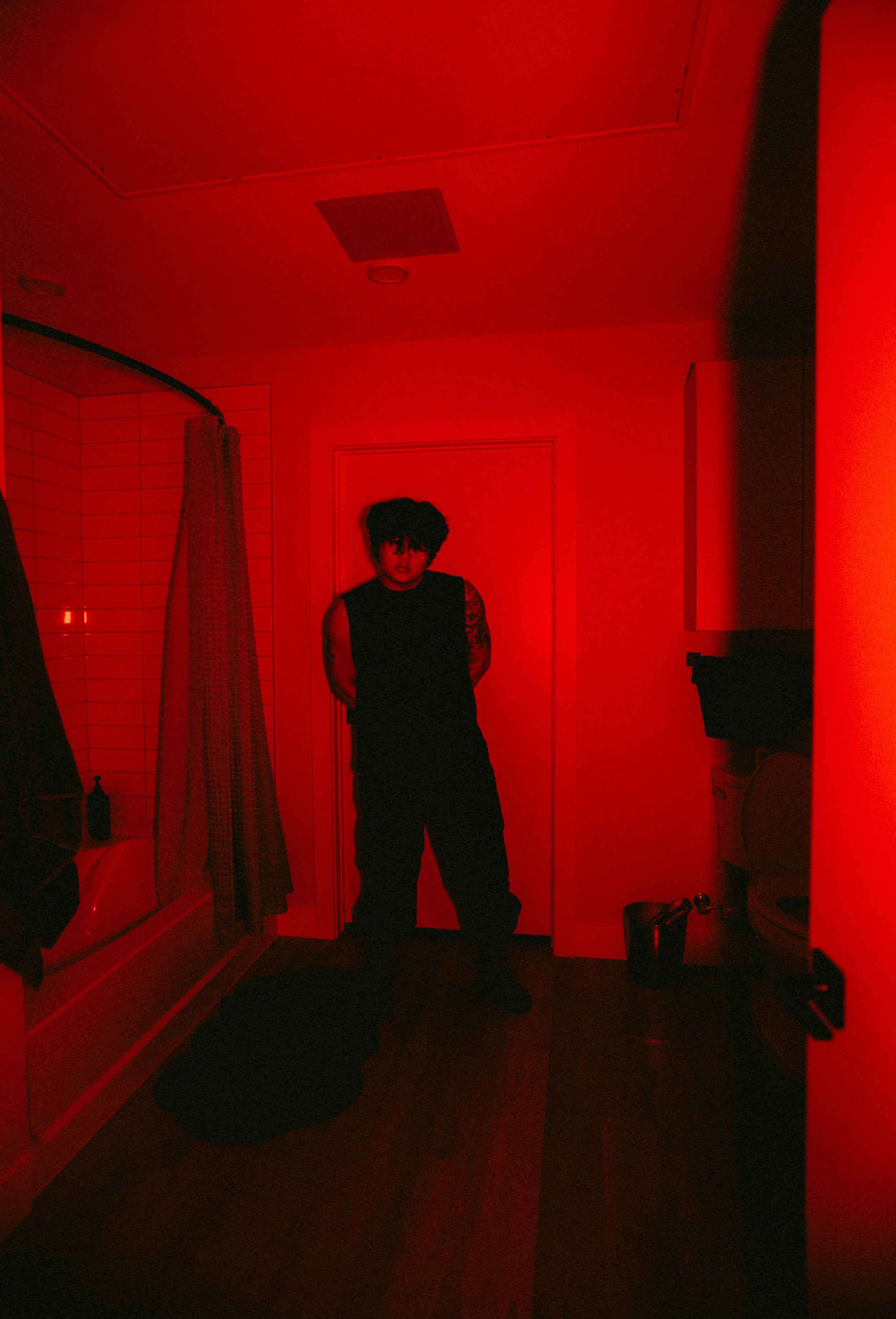 A person with curly hair standing in a dimly lit room illuminated by red light, facing the camera with hands behind their back. The room appears to be a bedroom or bathroom with tiled walls, a shower curtain, a closed door, and a computer monitor on 