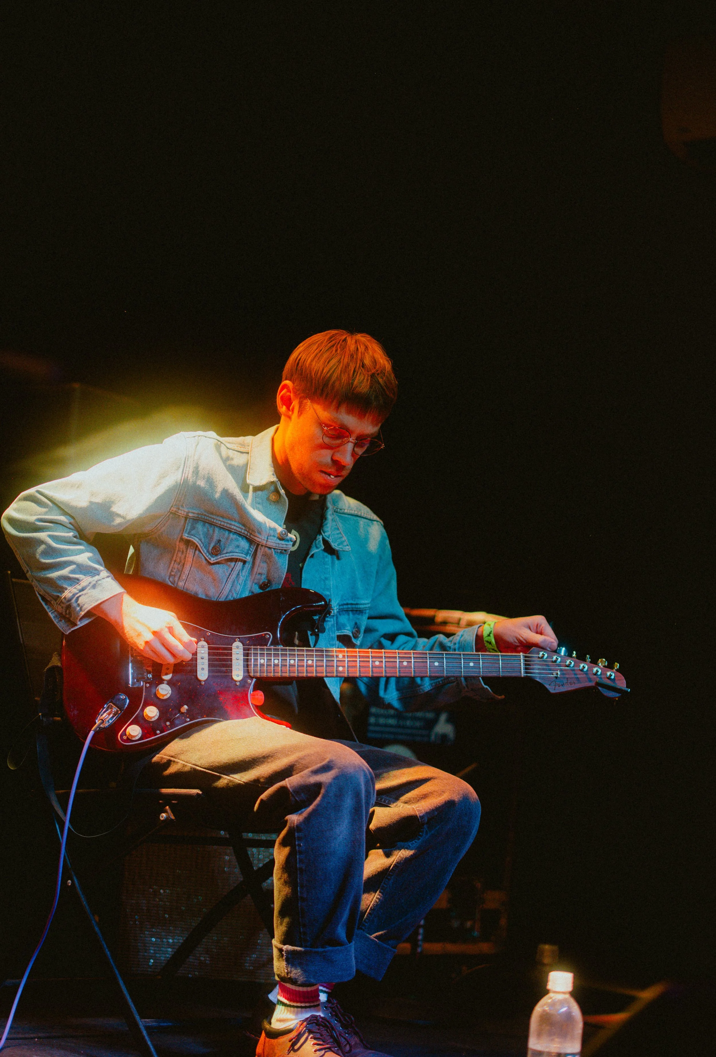 A young man playing an electric guitar on stage. He is wearing glasses, a denim jacket, and beige pants. There is a water bottle on the floor near his feet.