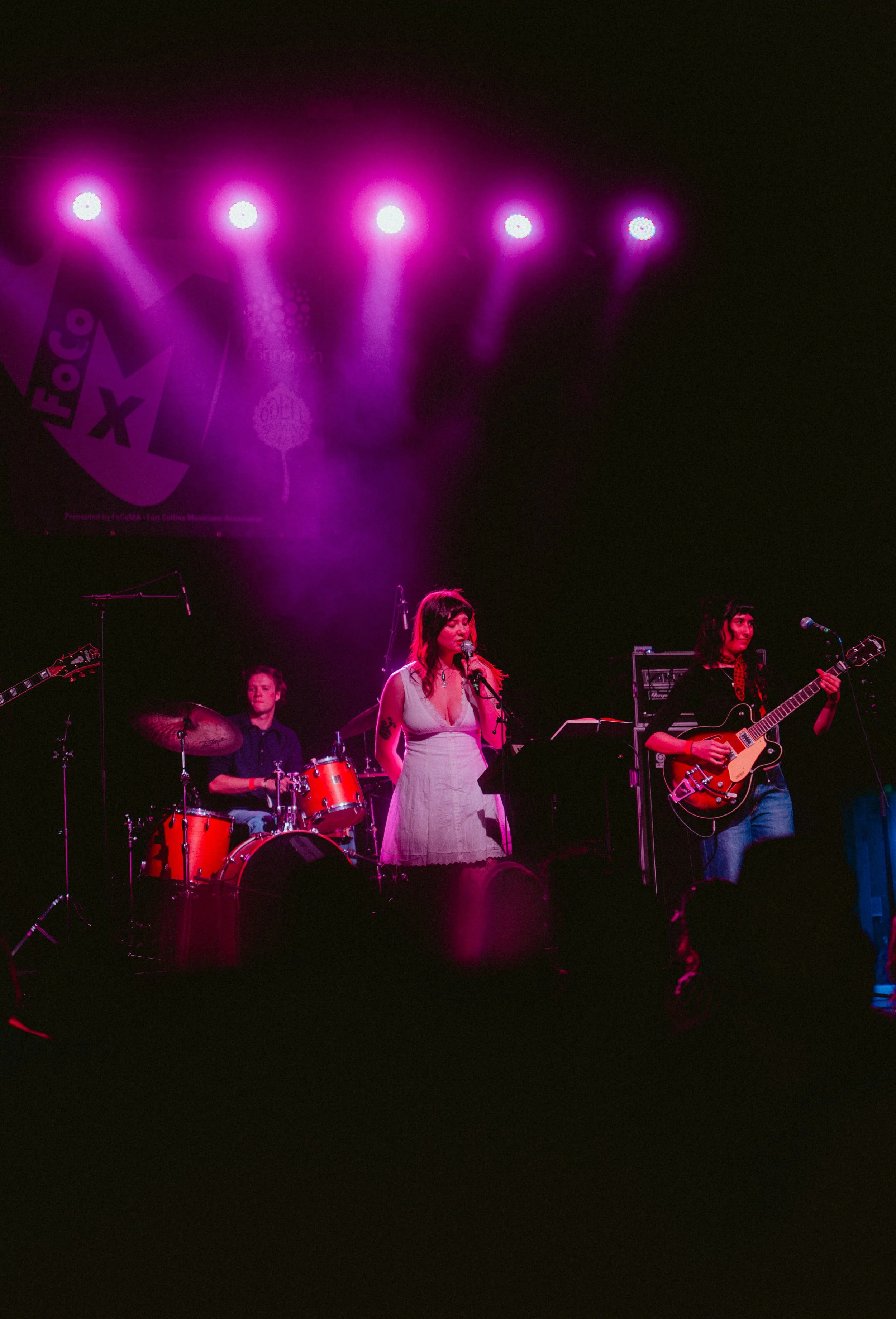 A band performing live on stage with pink lighting, featuring a drummer, a singer, and a guitarist.