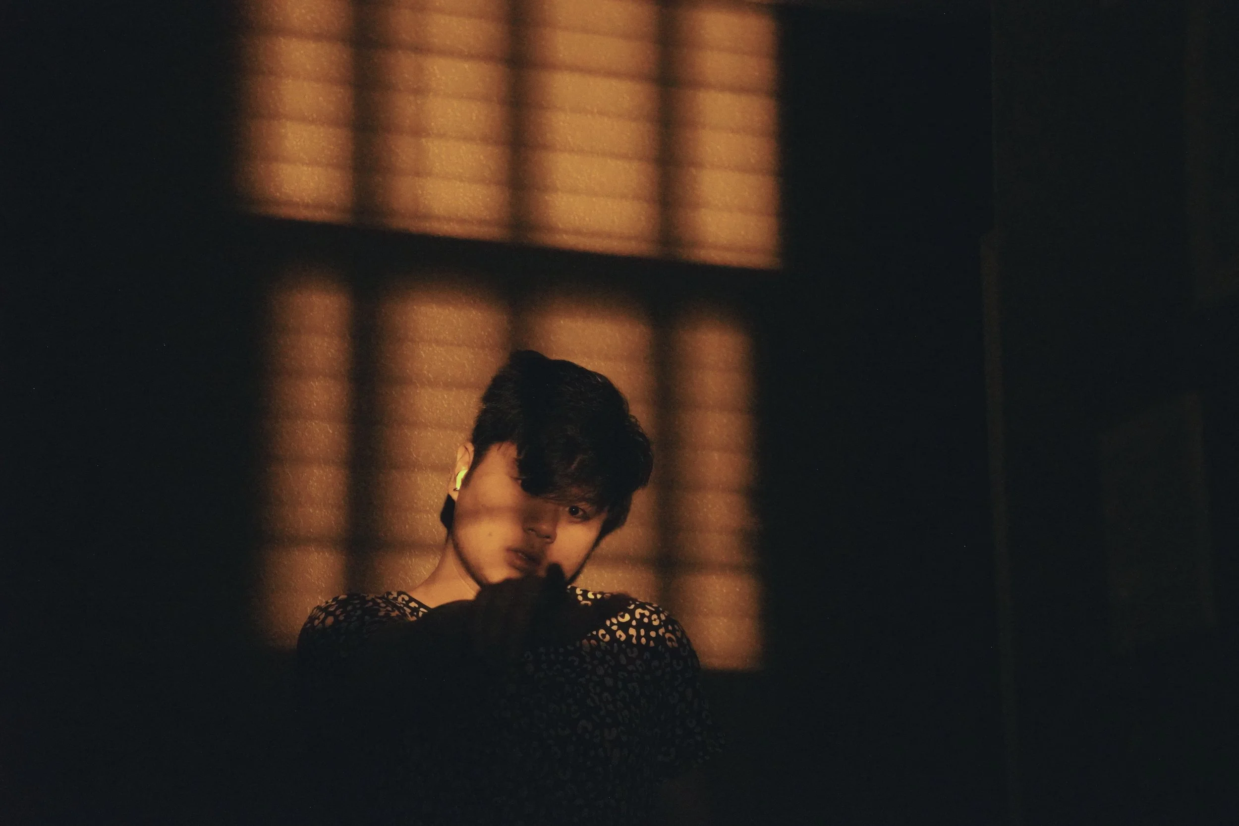 A person with short dark hair, wearing a patterned top, is standing with a serious expression. The background features a window with horizontal blinds casting warm, striped light onto the wall and floor, in a dimly lit room.