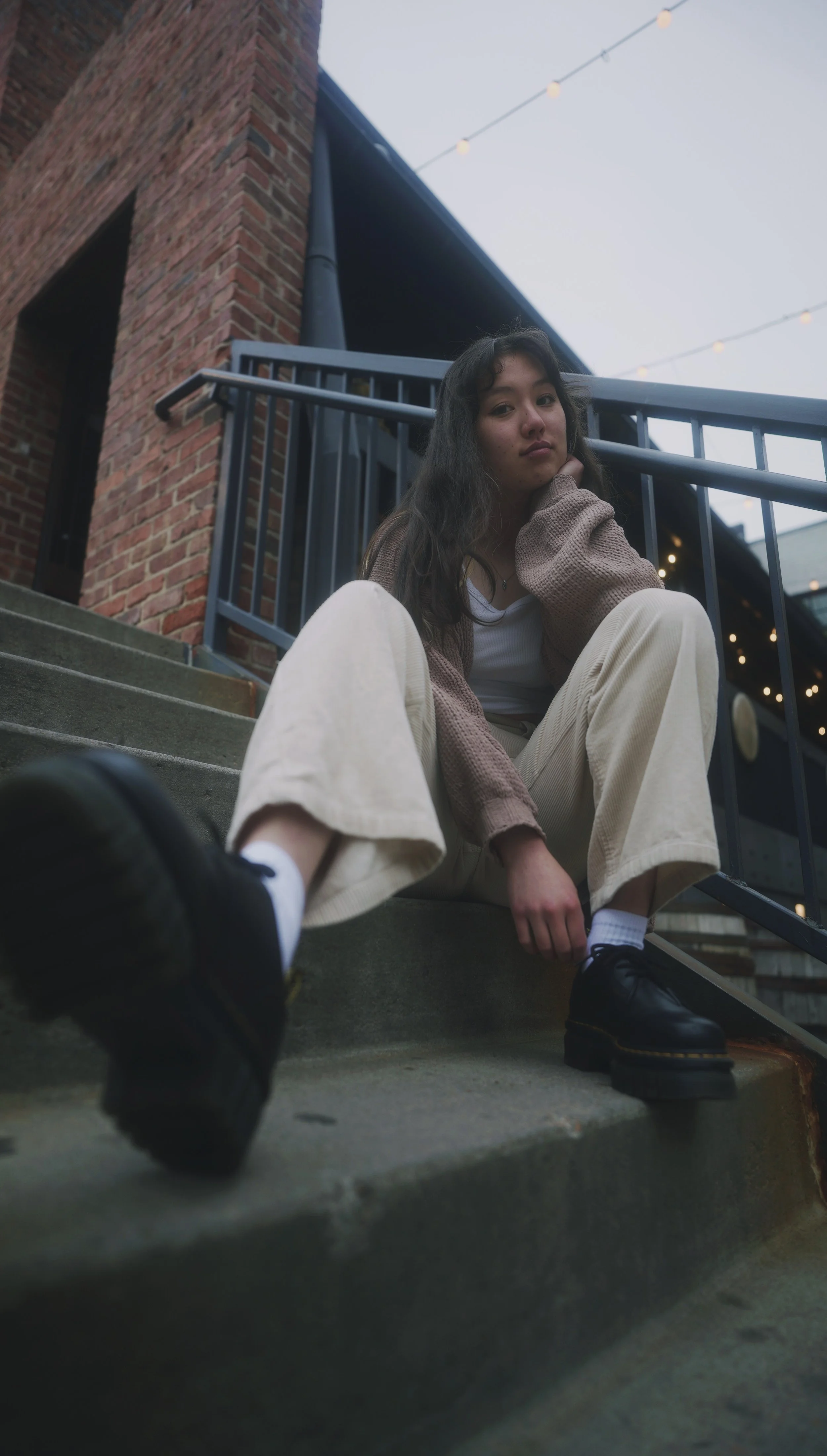 A young woman with long dark hair sitting on concrete stairs outside a brick building, looking at the camera with a contemplative expression, wearing beige pants, a white shirt, a brown cardigan, and black shoes.