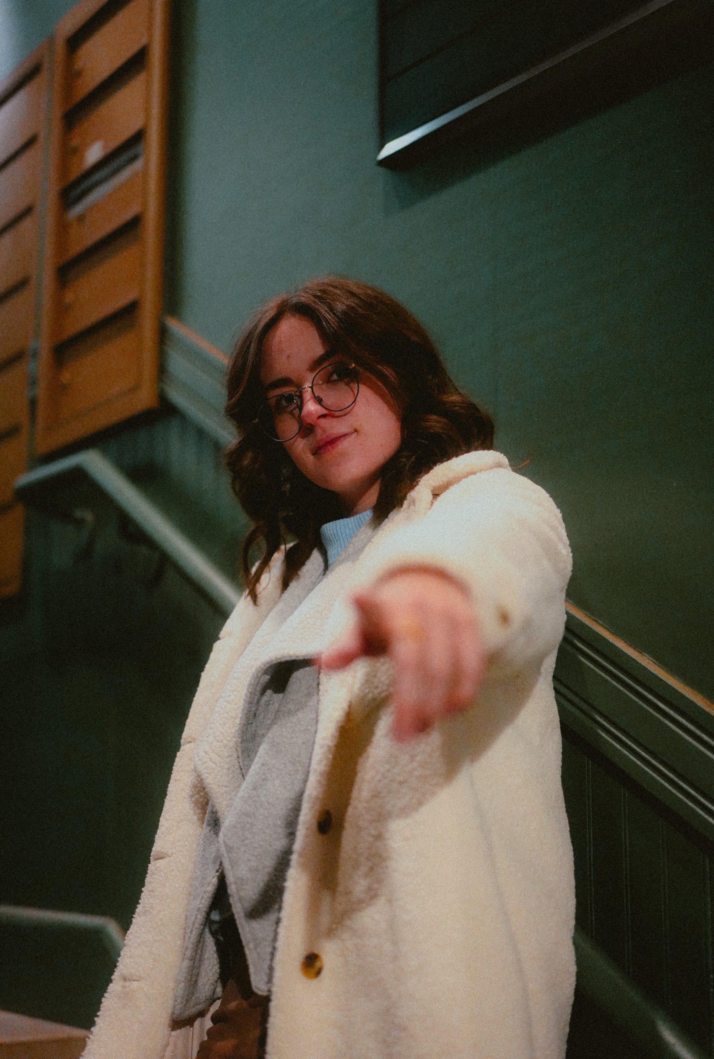 A woman with brown hair and glasses pointing towards the camera while standing in front of a green wall with wooden and black cabinets.