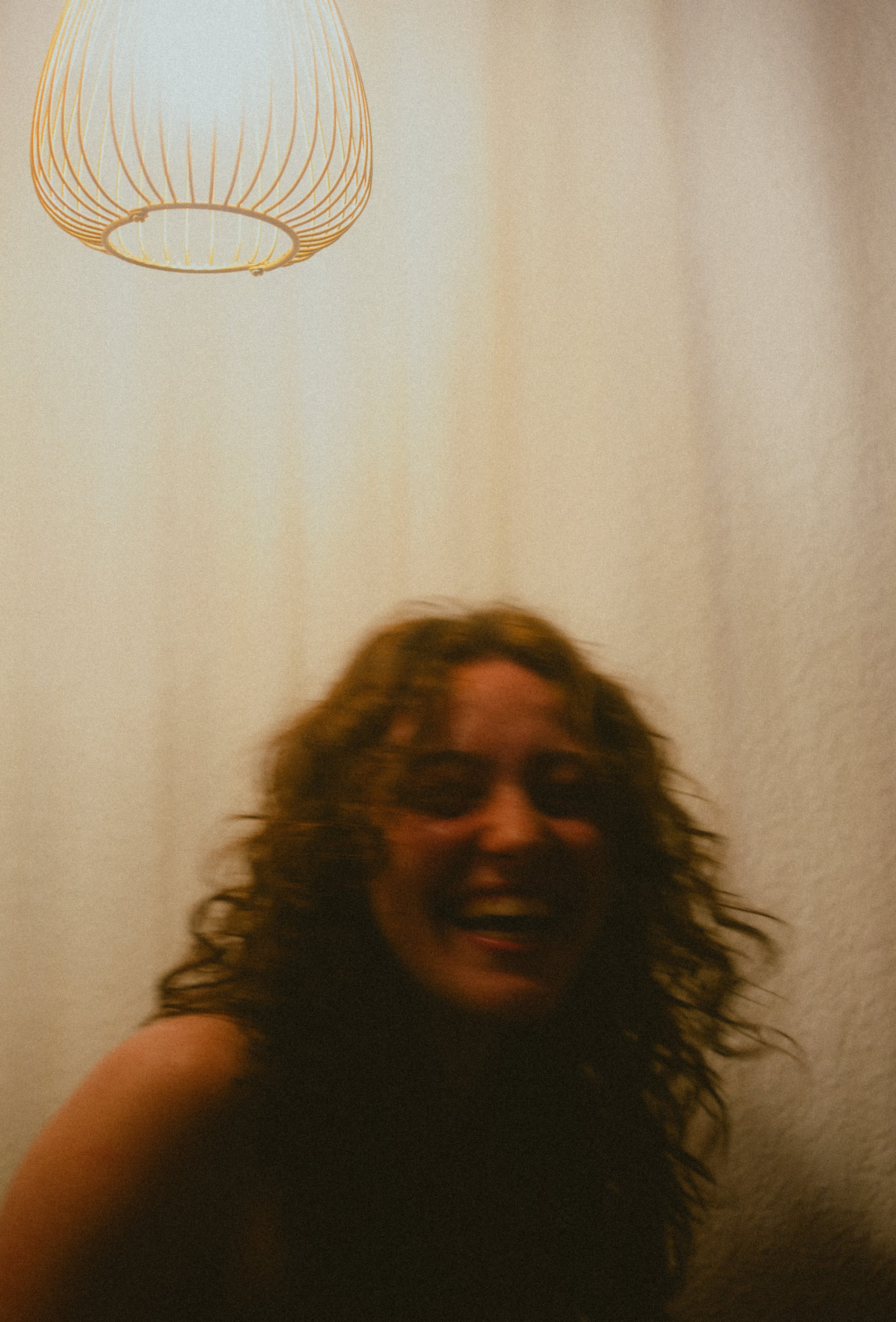 A person with long curly hair and sunglasses is smiling and taking a selfie, with a light-colored wall and a hanging lamp in the background.