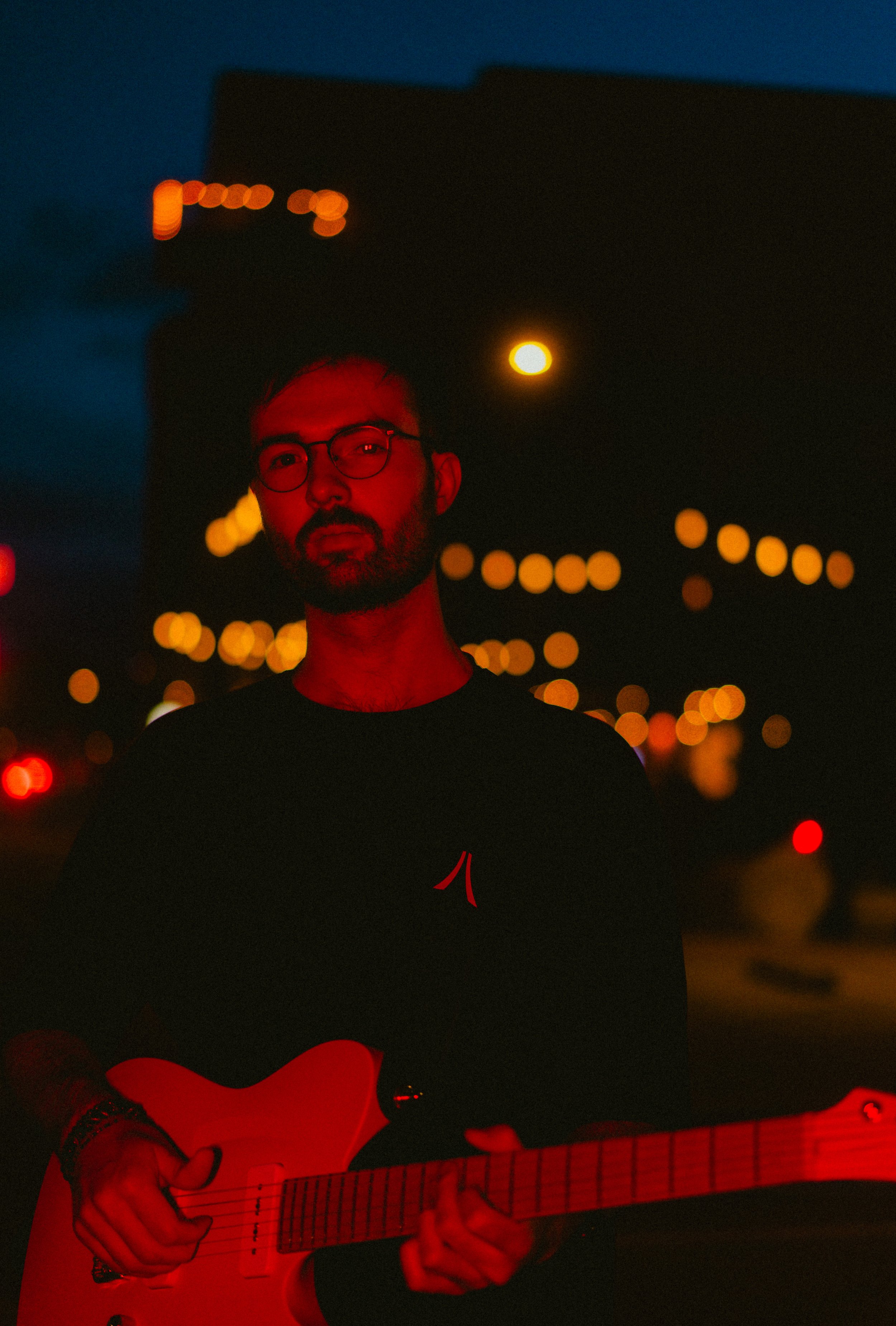 A man with glasses and a beard playing an acoustic guitar under red and yellow streetlights at night.