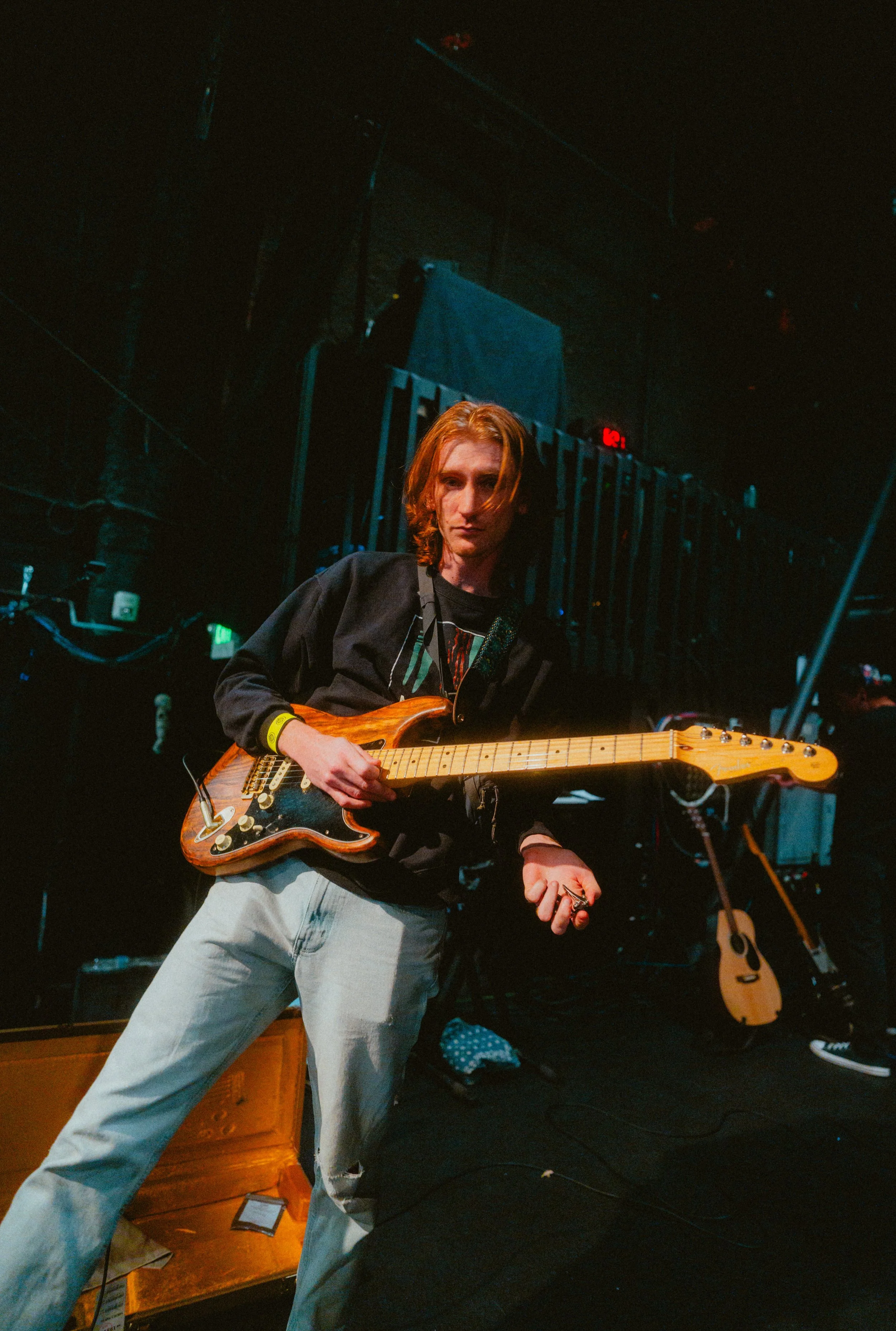 A young man with long, red hair is playing an electric guitar on stage. He is wearing a black jacket, light-colored pants, and a yellow wristband, with a focused expression. The stage has various musical instruments and equipment, and a second acoust