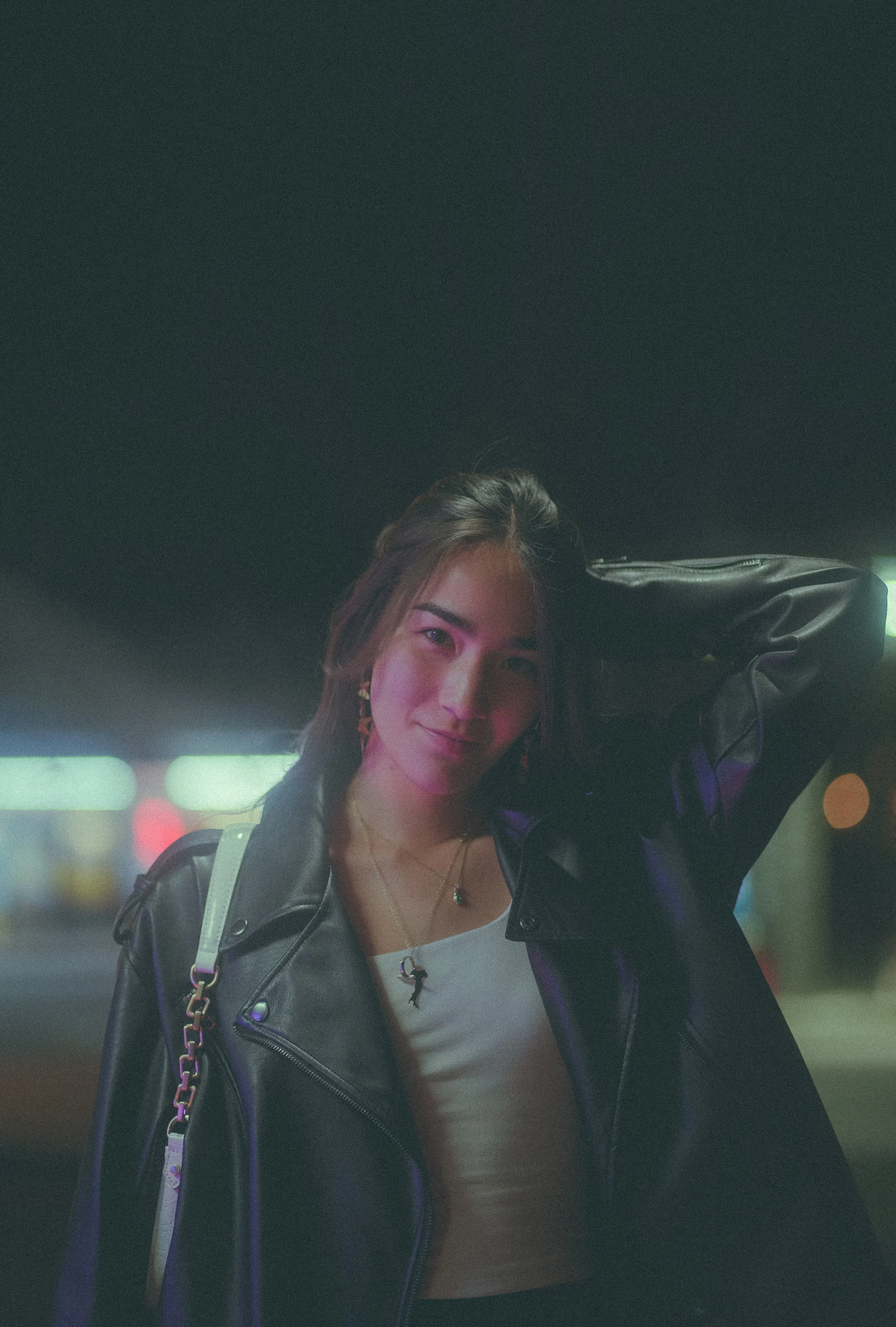 A young woman with brown hair smiling, wearing a white shirt and black leather jacket, standing outdoors at night with blurred colorful lights in the background.