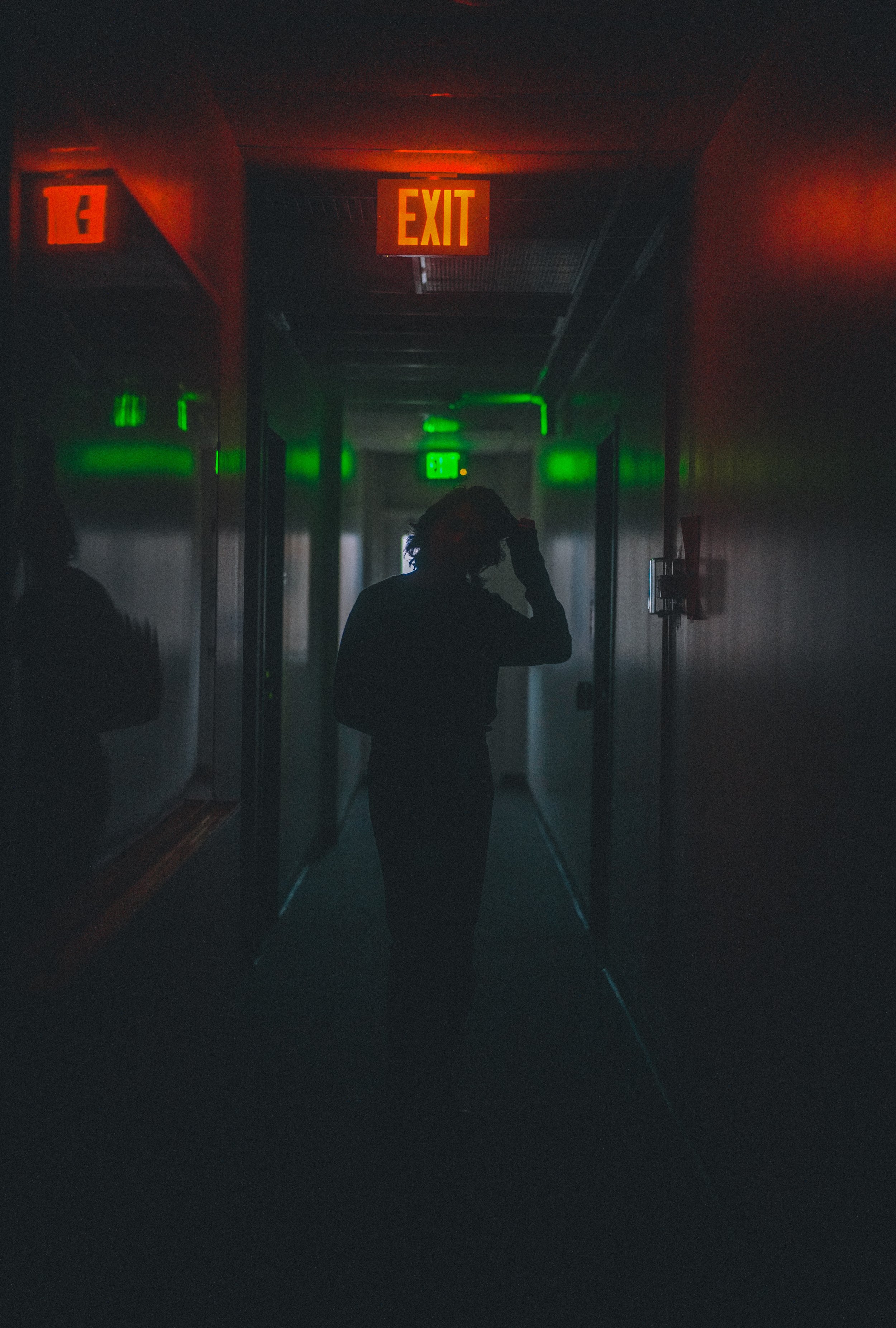 Dark corridor with green and red lights, a person silhouetted in the center, and illuminated exit signs at the end of the hallway.
