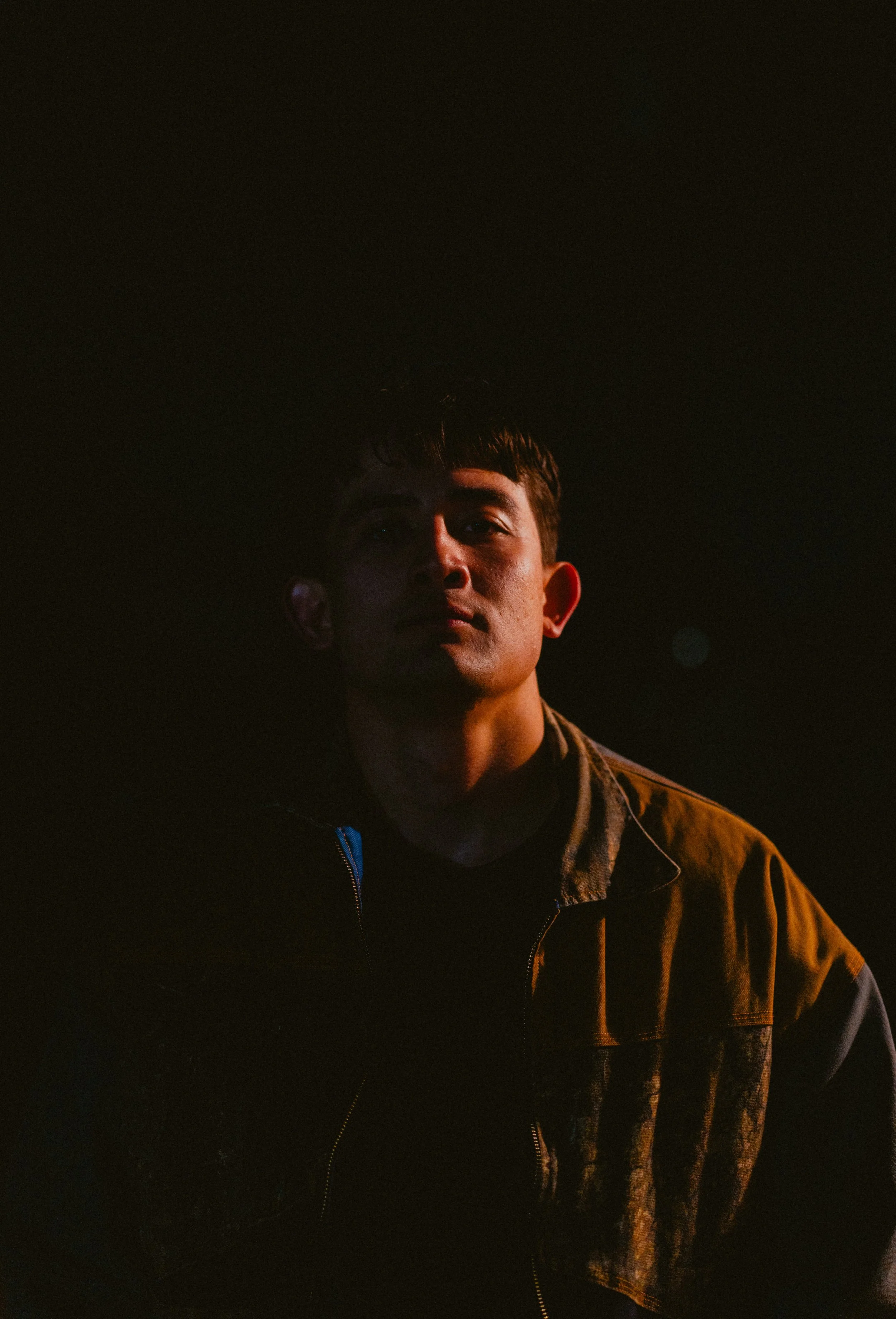 A young man with short dark hair against a dark background, wearing a brown jacket with a camouflage pattern on the shoulders.
