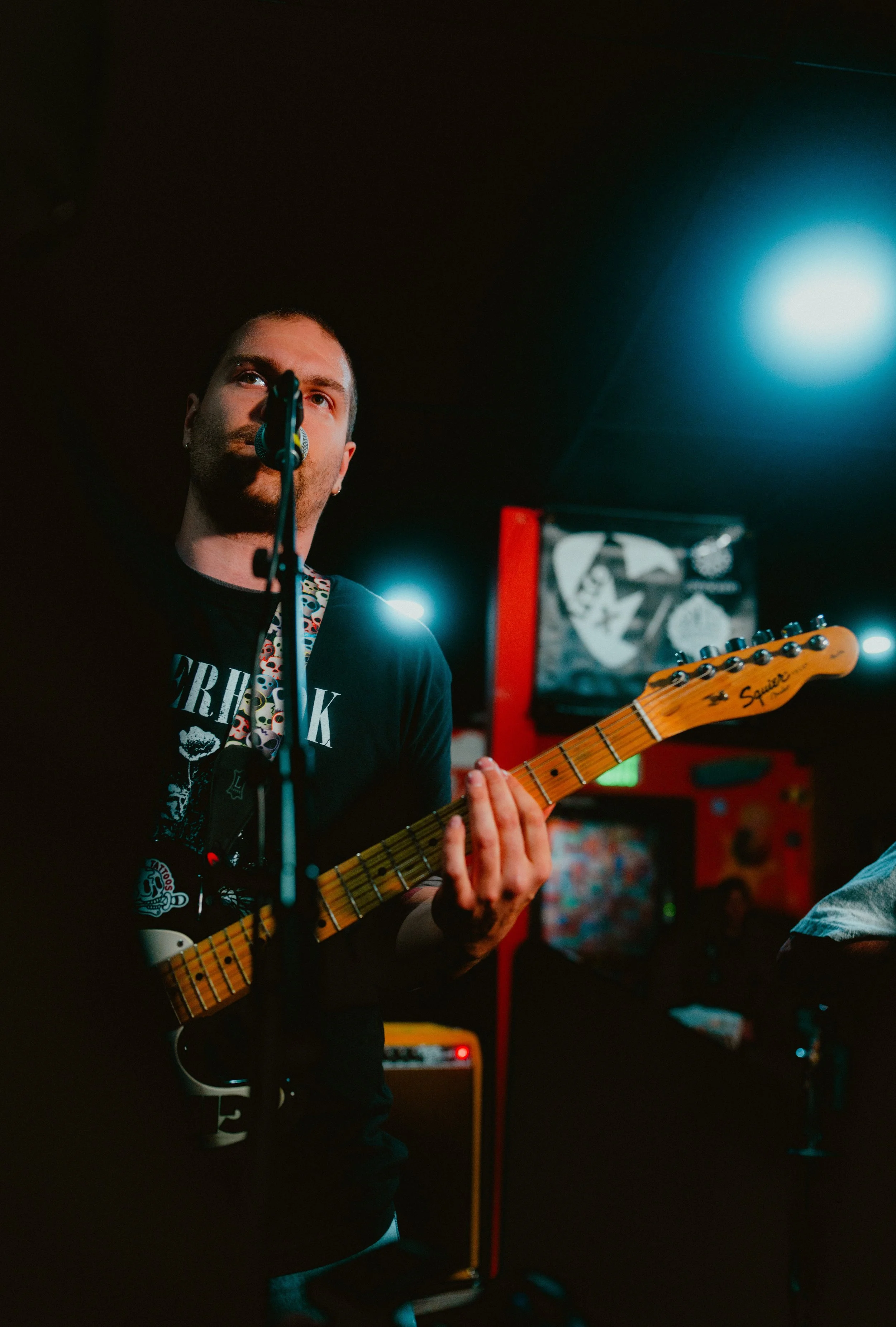 A man playing an electric guitar singing into a microphone in a dimly lit room with colorful posters and equipment in the background.