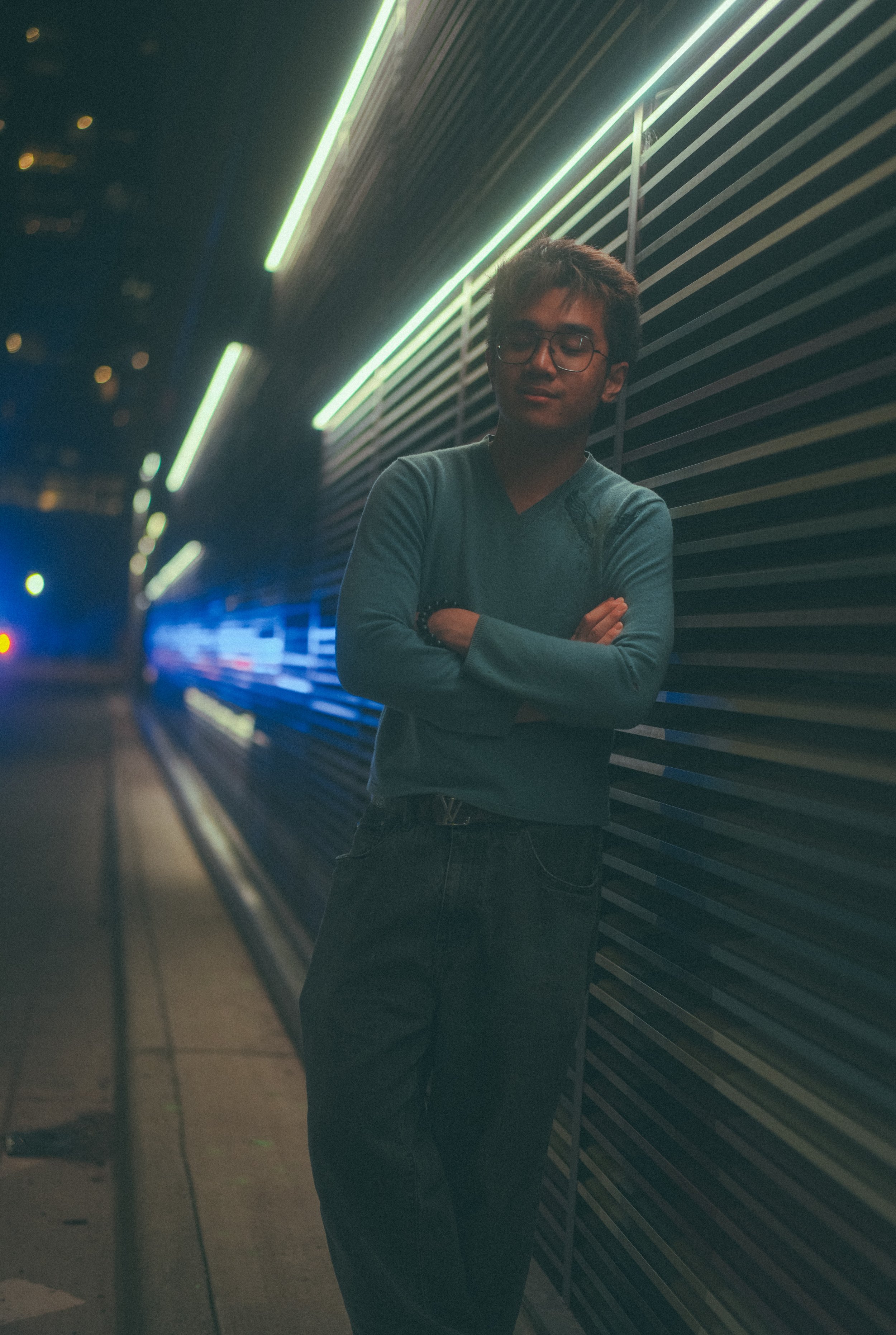 A young man with glasses and dark hair stands with arms crossed in front of a dark wall with horizontal lines and neon lights, at night.