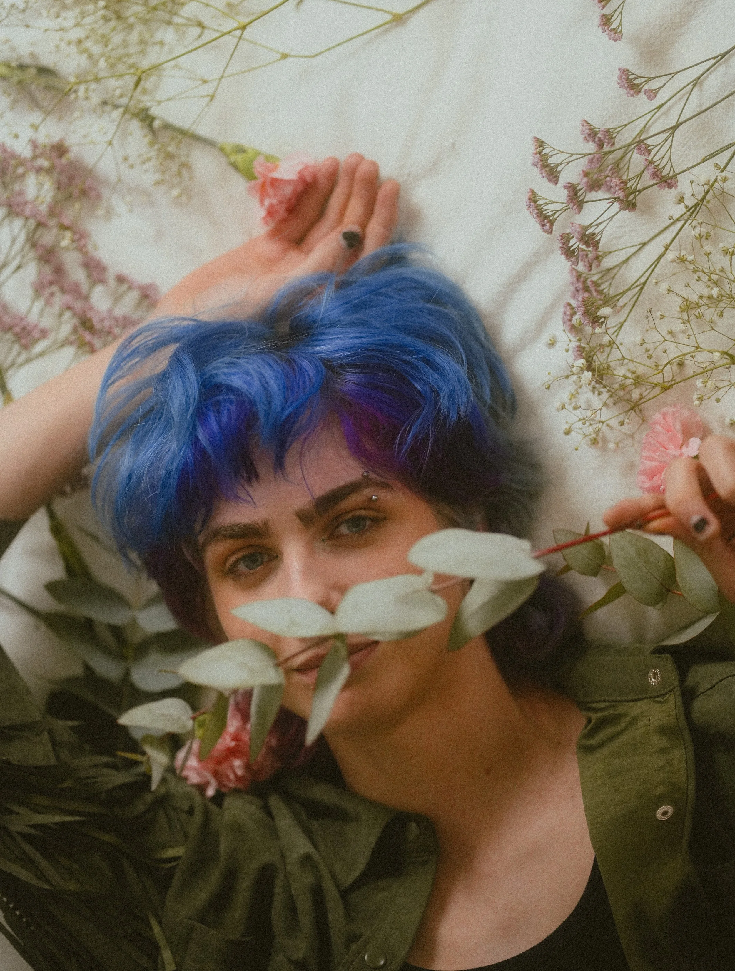 A person with blue and purple hair lying among pink and white flowers and green leaves, wearing a green jacket and a black top.