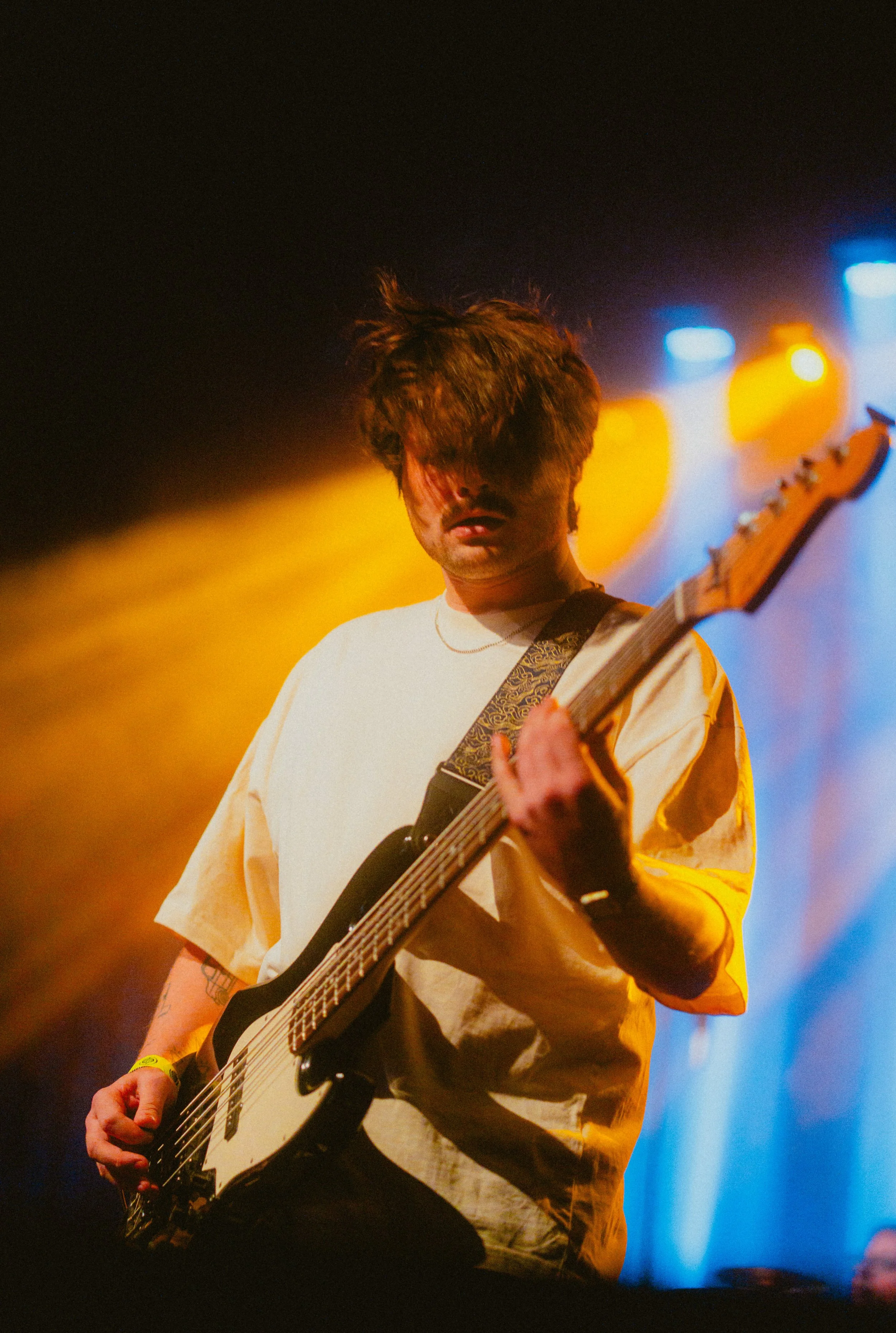 A young man with messy brown hair playing an electric guitar on a stage with colorful blue and orange stage lights.