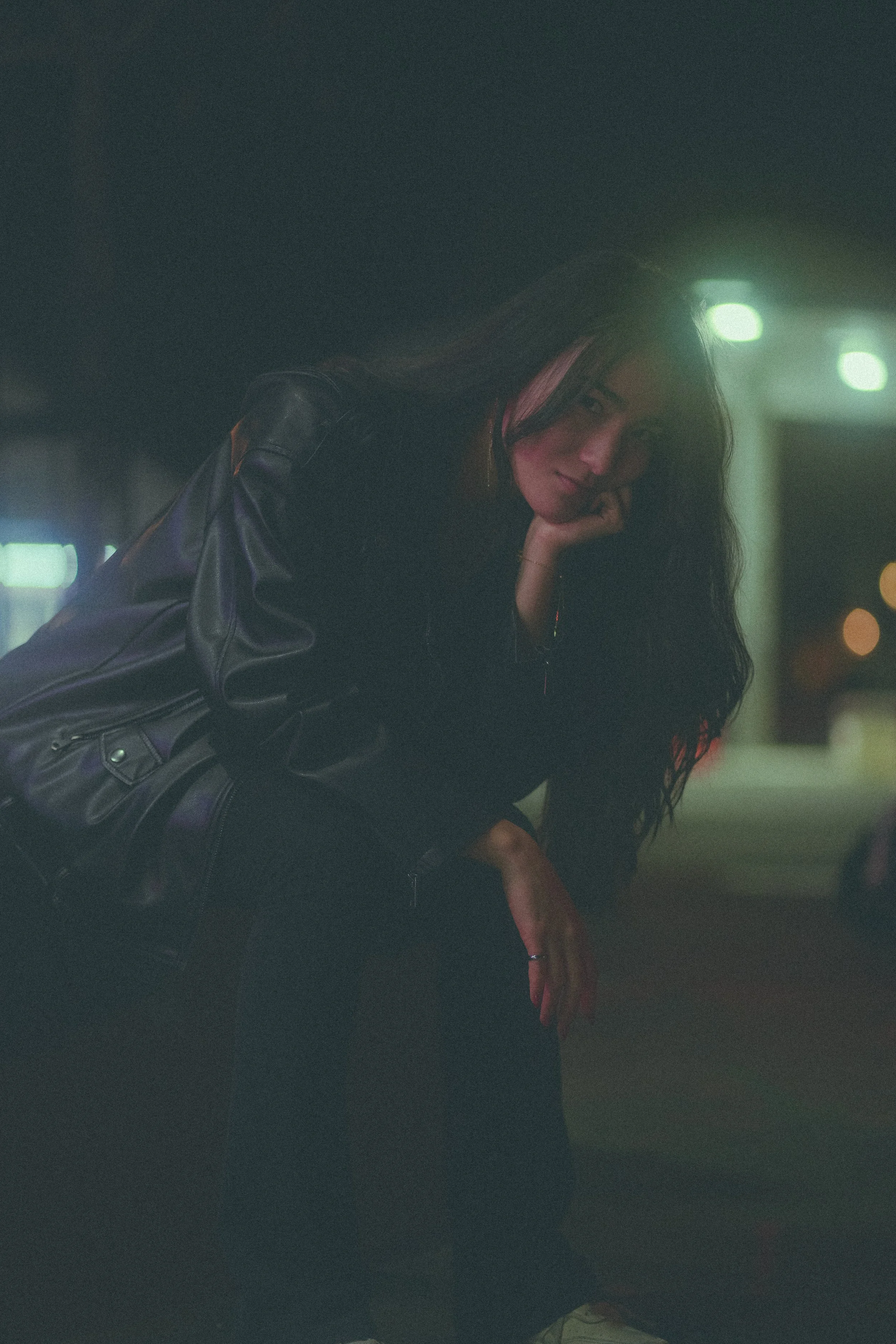 A woman with long dark hair, wearing a leather jacket, resting her chin on her hand, looking at the camera in a dimly lit setting with blurred lights in the background.