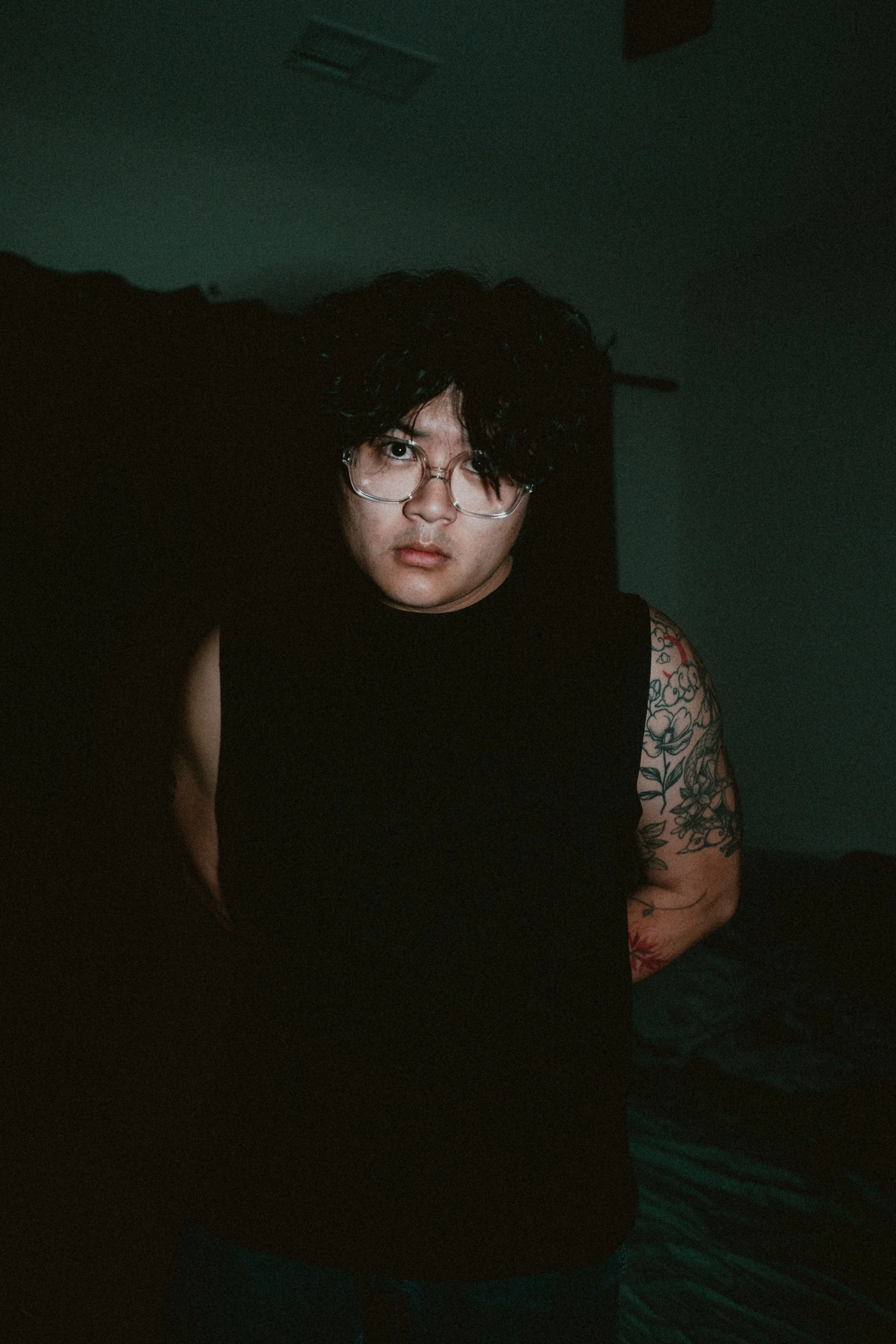 A young man with curly black hair, glasses, and tattoos on his arm, wearing a sleeveless black shirt, sitting in a dimly lit room with a teal wall.