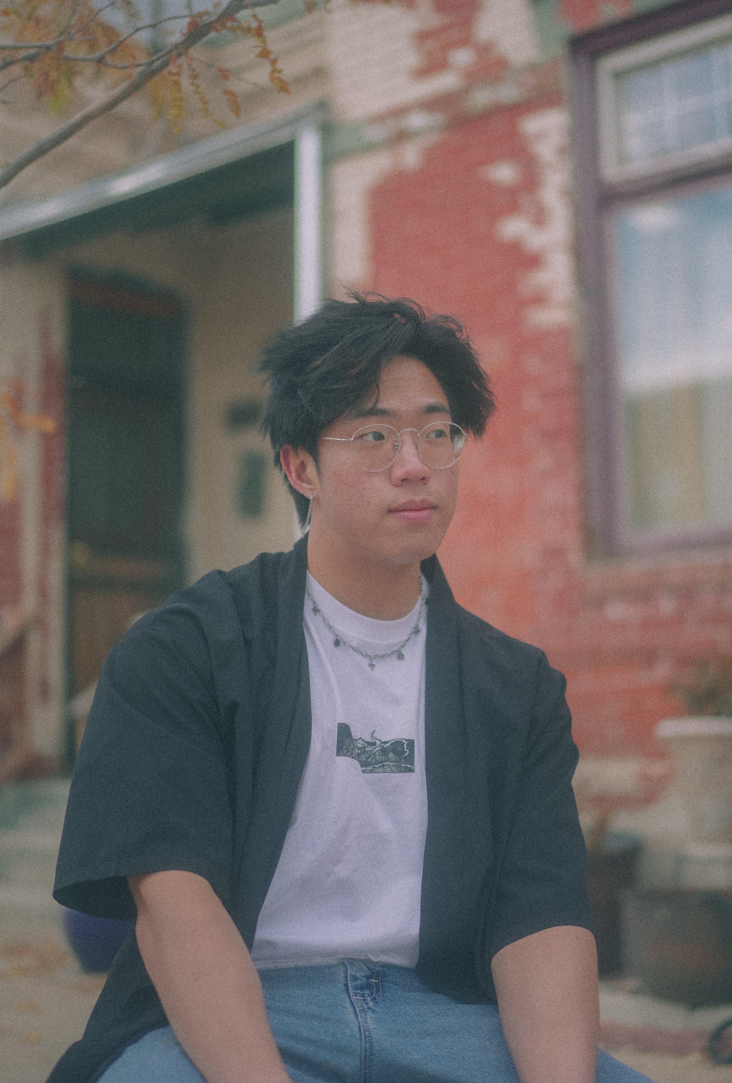 A young man with black hair, glasses, and earrings sitting outdoors with a blurred brick building in the background.