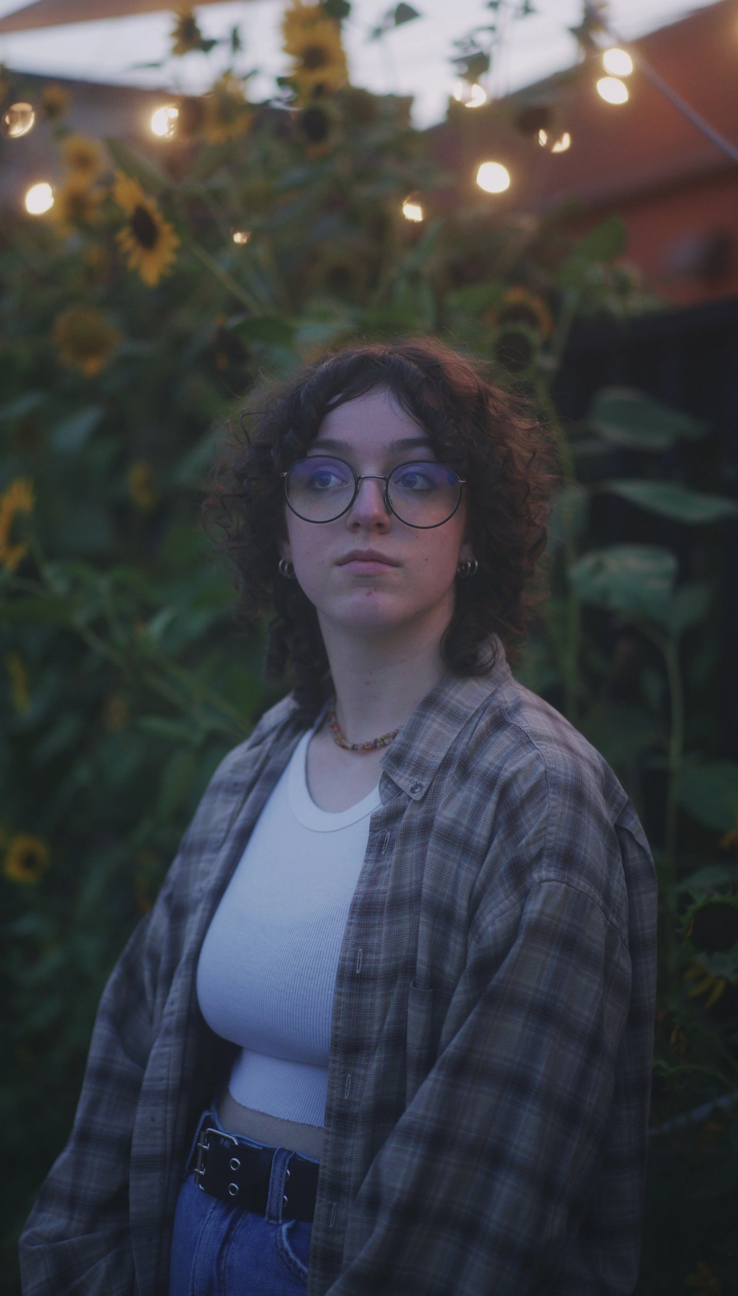 A young person with curly hair and round glasses standing outdoors among sunflowers, wearing a white crop top, a plaid shirt, and a beaded necklace, with string lights above during twilight.