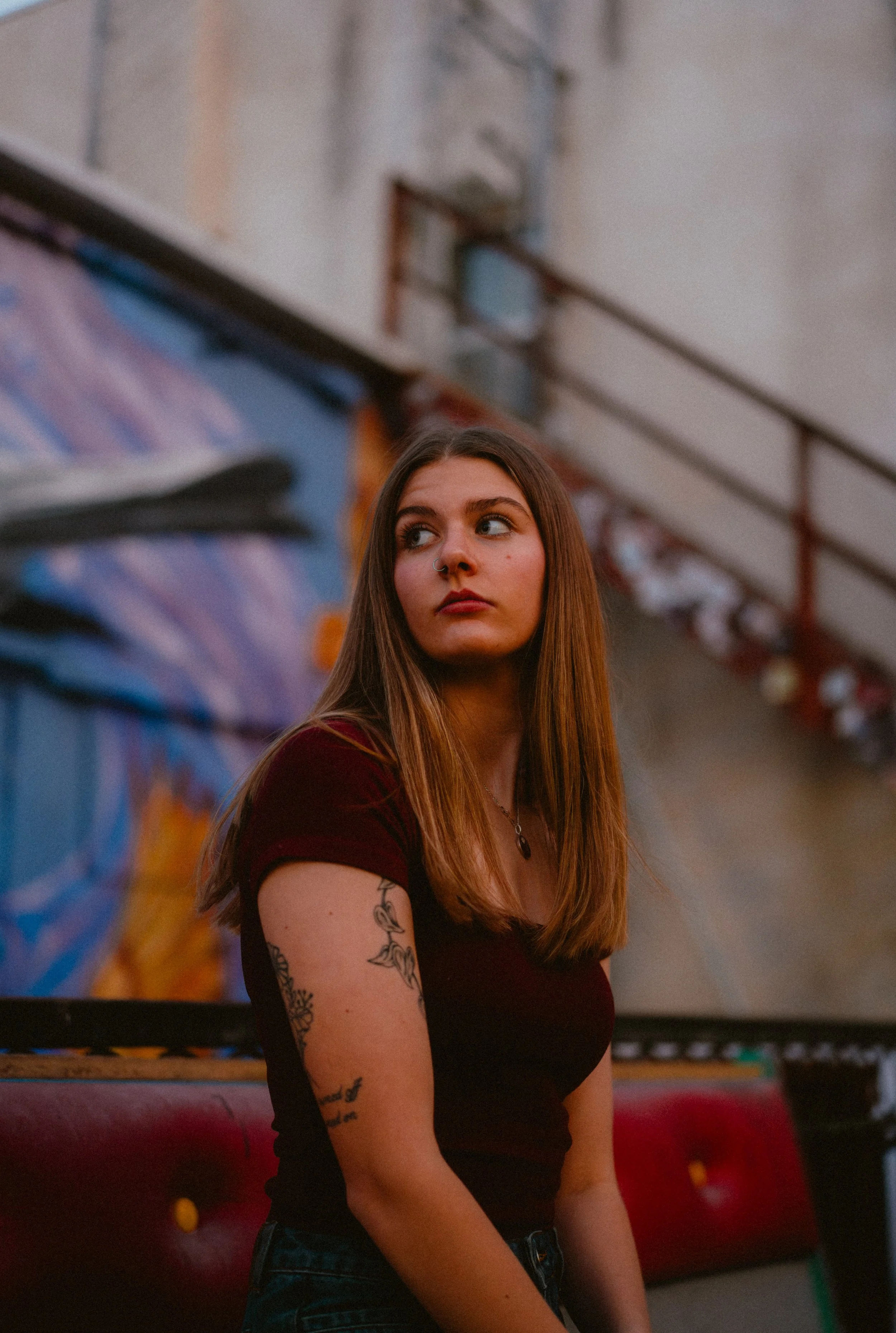 A young woman with long brown hair, tattoos on her arms, wearing a black shirt, standing in front of a colorful mural in an indoor skate park or similar setting.