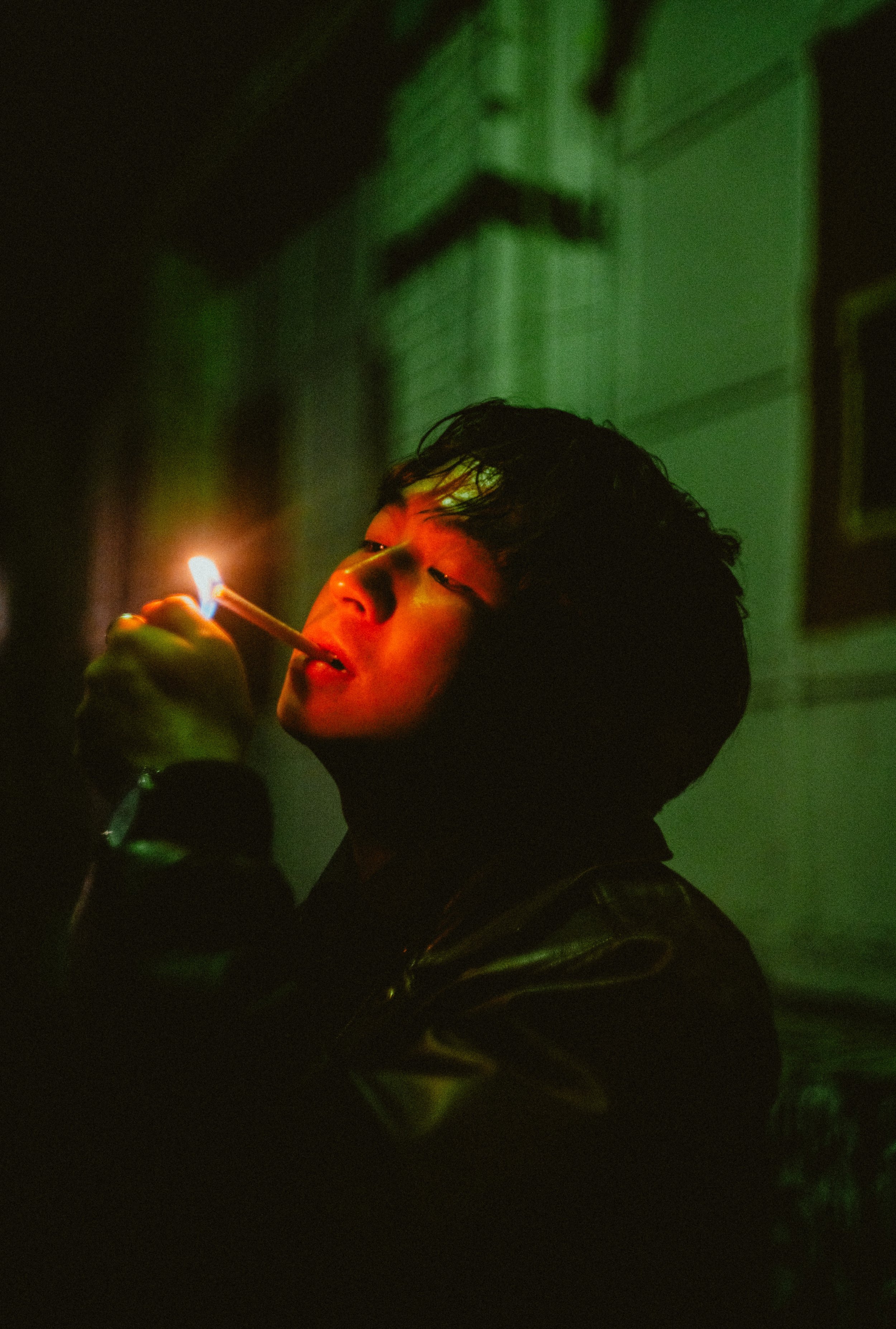A young person with dark hair is holding a lit match close to their face in a dimly lit room with green lighting, creating a dramatic effect.