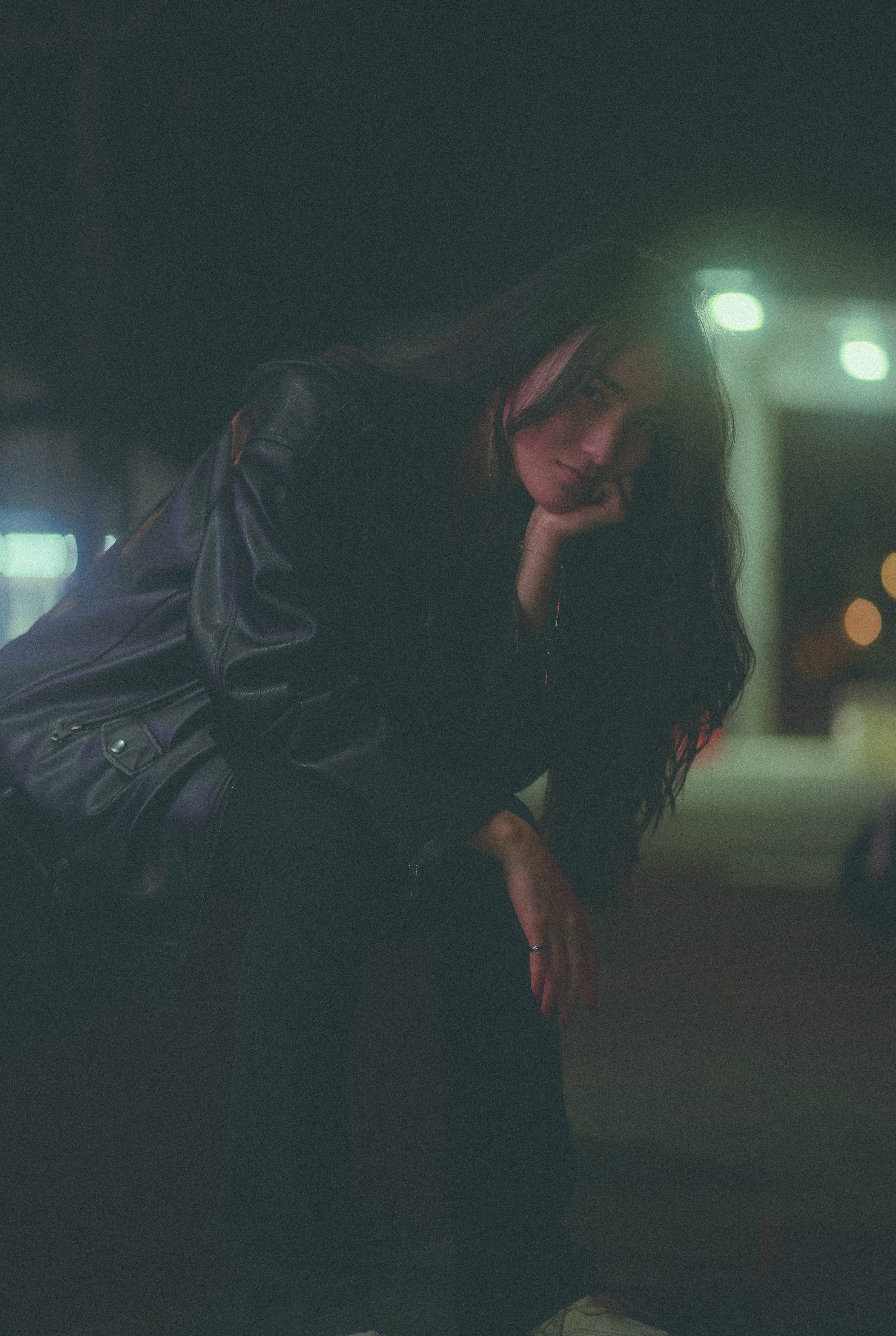 Young woman with long dark hair and tattoos, wearing a black leather jacket, sitting and resting her face on her hand in a dimly lit indoor environment with green and orange lights.