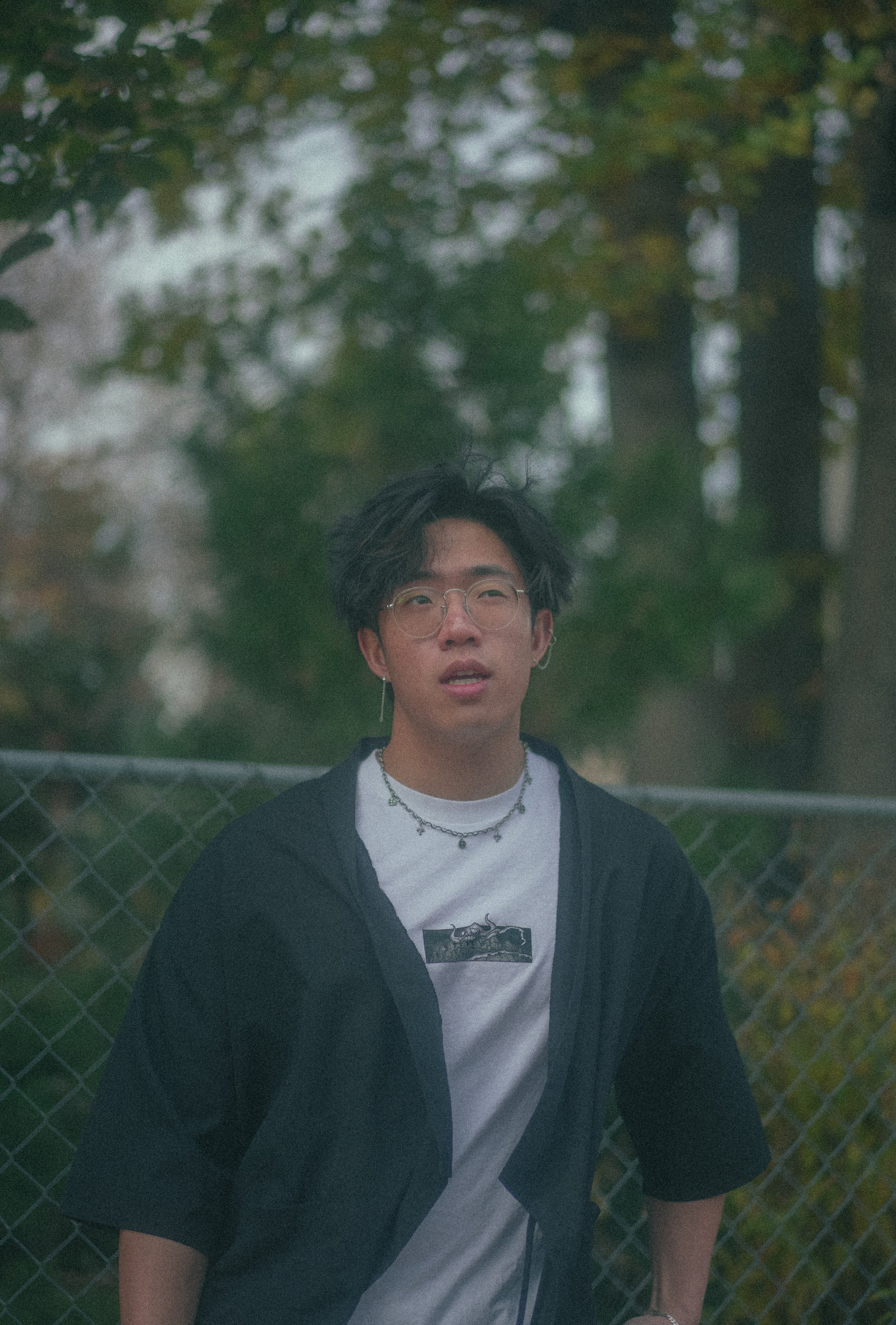 A young man with glasses, wearing a white graphic T-shirt, a black jacket, and jewelry, sitting outdoors in front of a chain-link fence with green trees in the background.