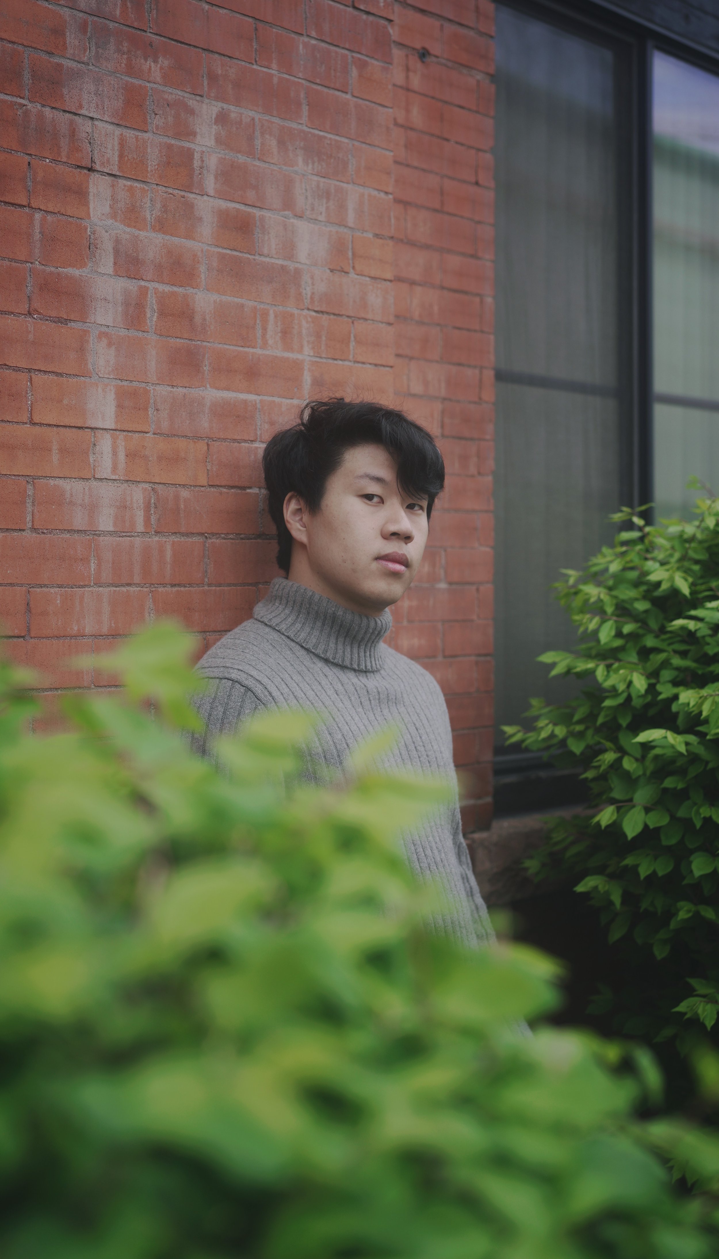 A young man with black hair wearing a gray turtleneck sweater leaning against a red brick wall, partially obscured by green bushes in the foreground.